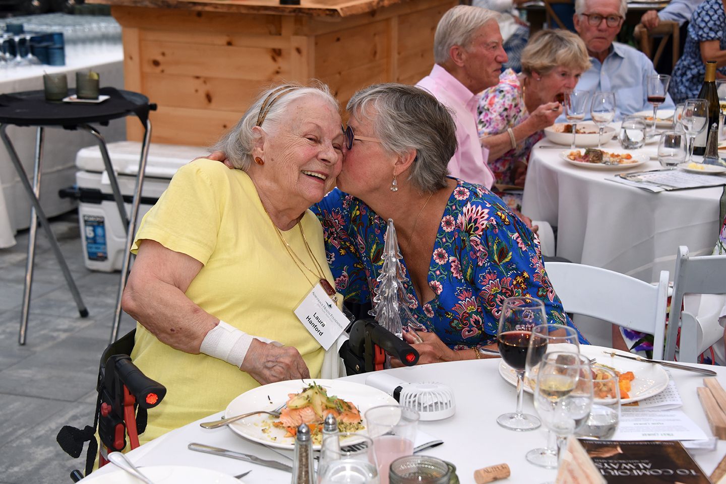 Two older women smiling, one kissing the other's cheek at a dinner table. People eat and chat at a restaurant.