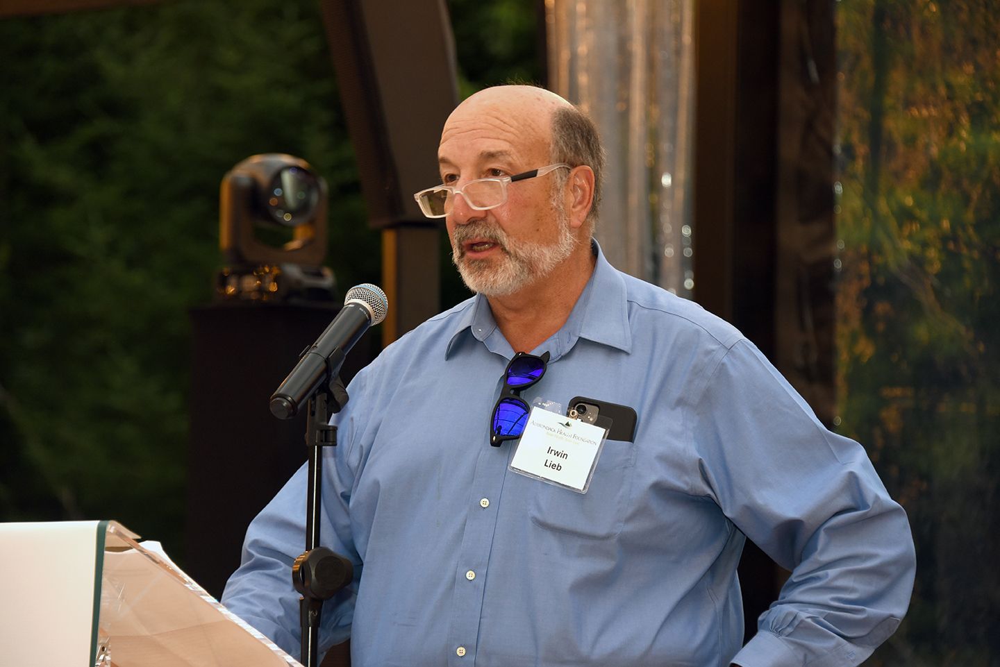 Man with glasses speaks into a microphone at an outdoor event; he wears a blue shirt and a name tag.