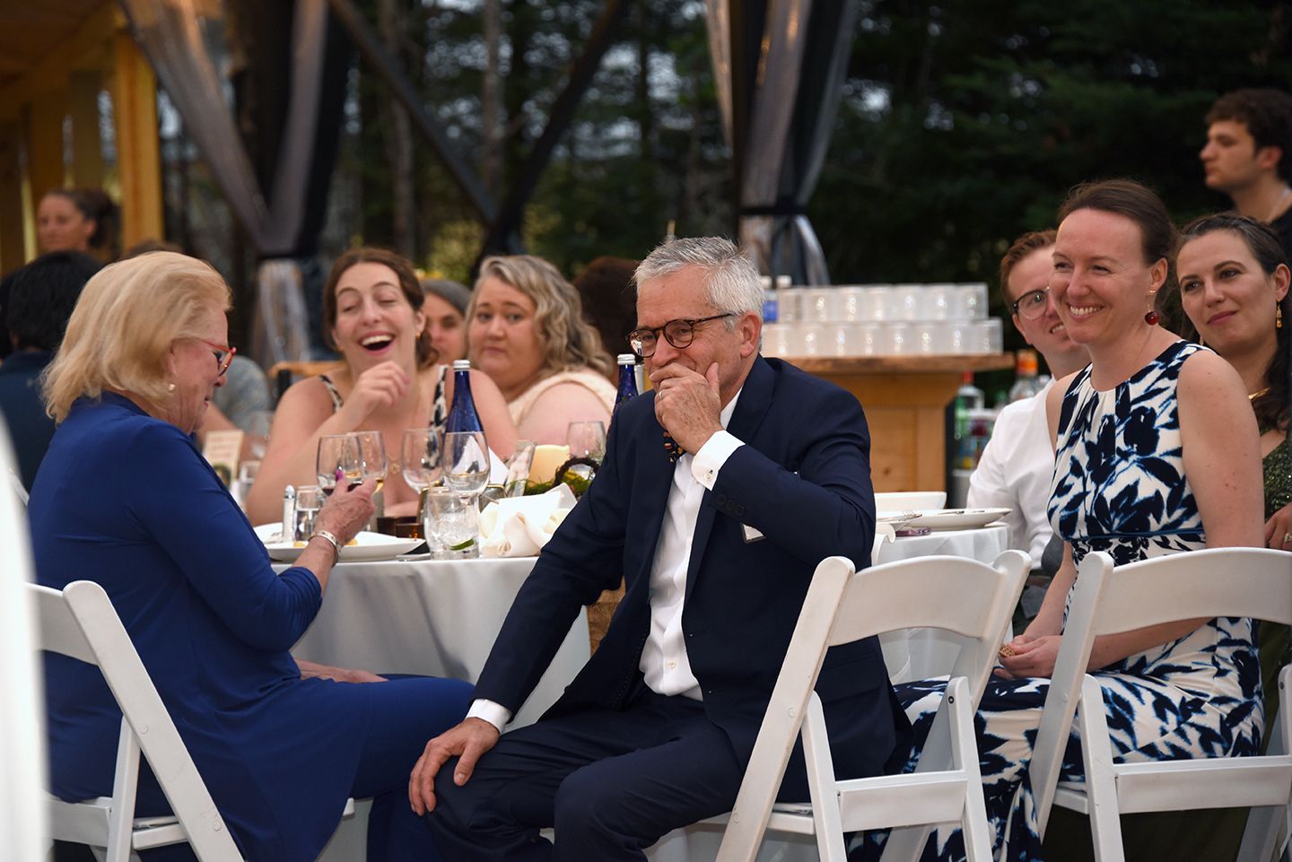 People seated at a table, at an outdoor event. One man in a blue suit gestures while speaking. Others laugh and smile.