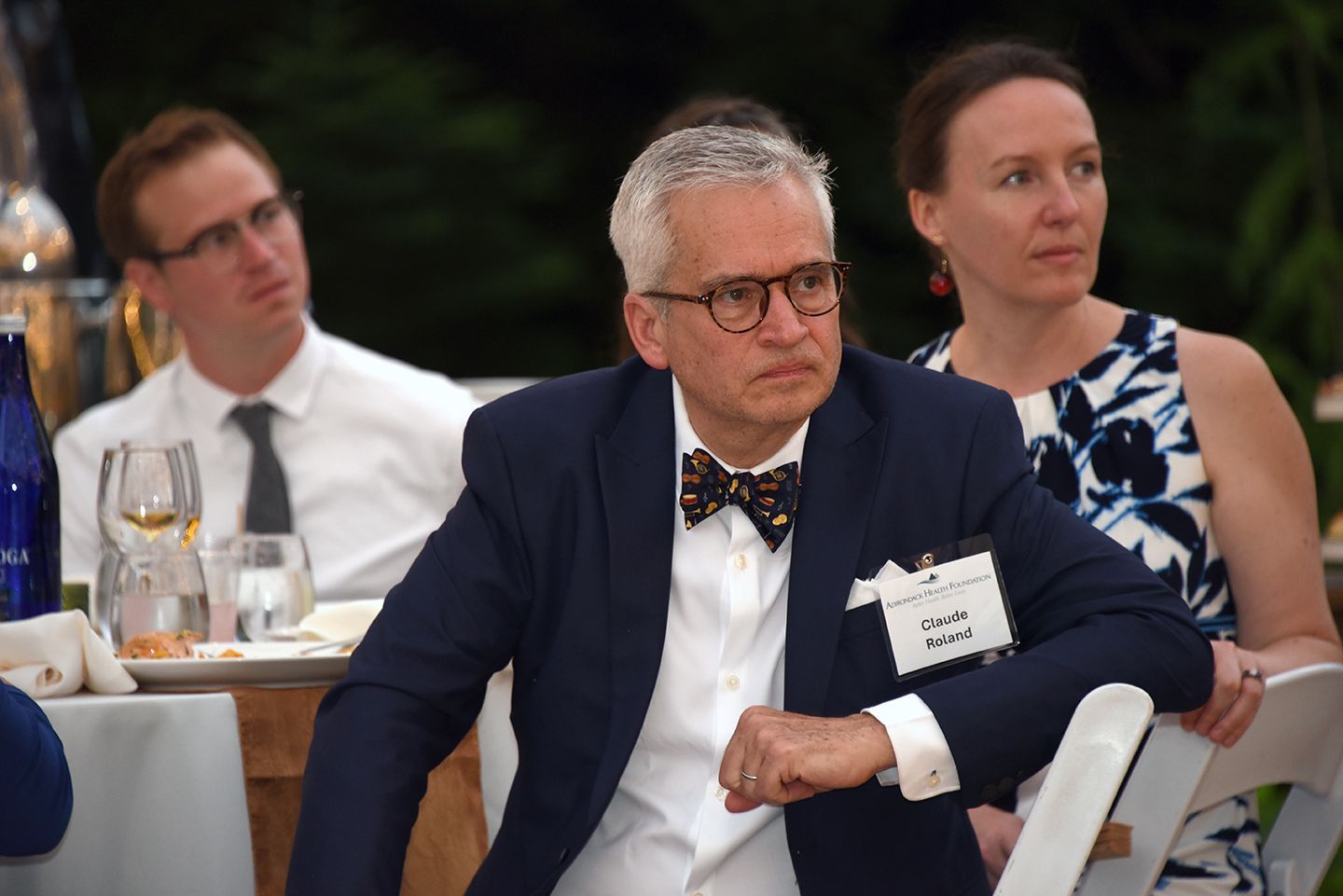 Man with gray hair, glasses, and bowtie at an outdoor event, looking to the side.