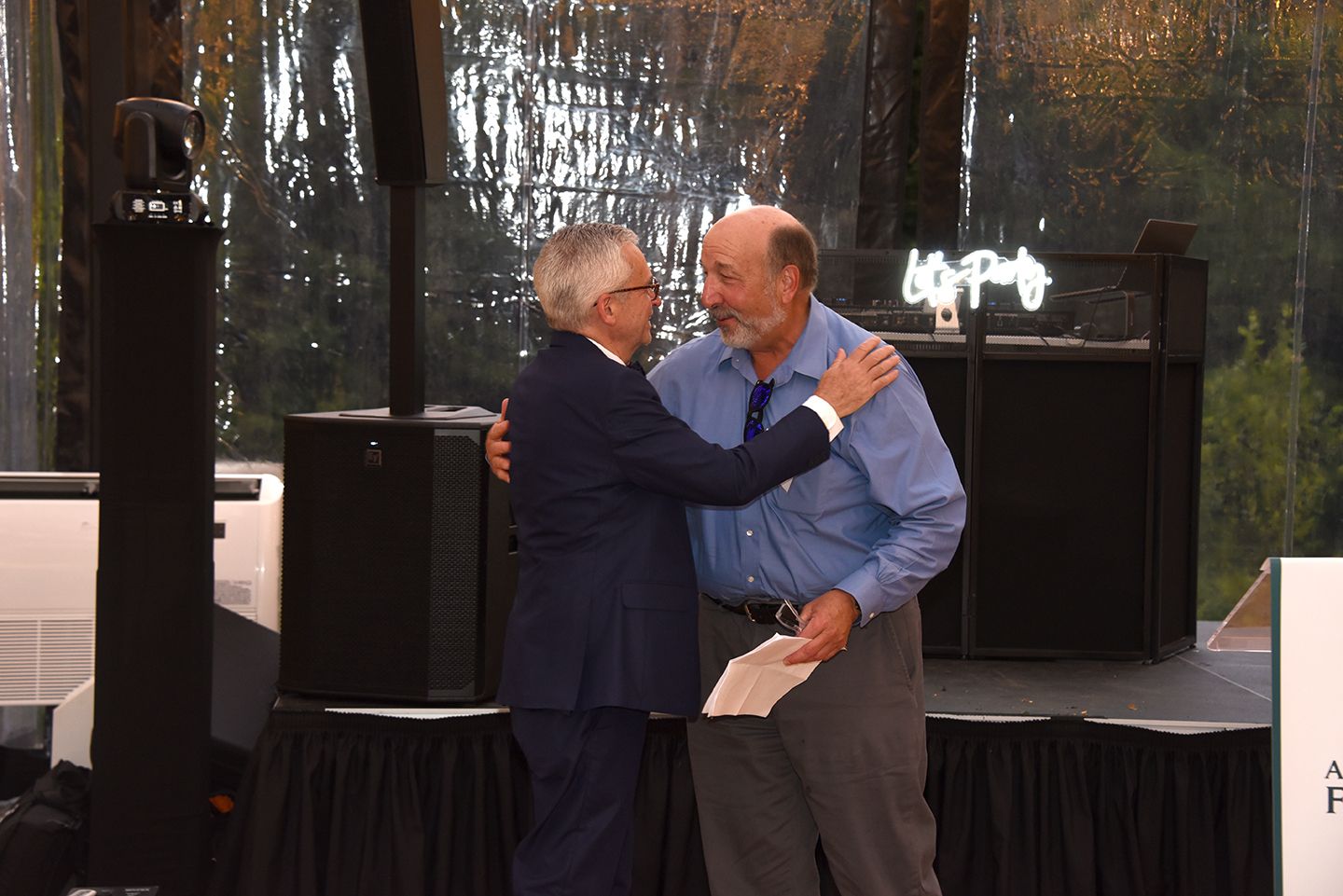 Two men embracing on a stage, one in a suit, the other in a blue shirt, possibly at an awards ceremony.