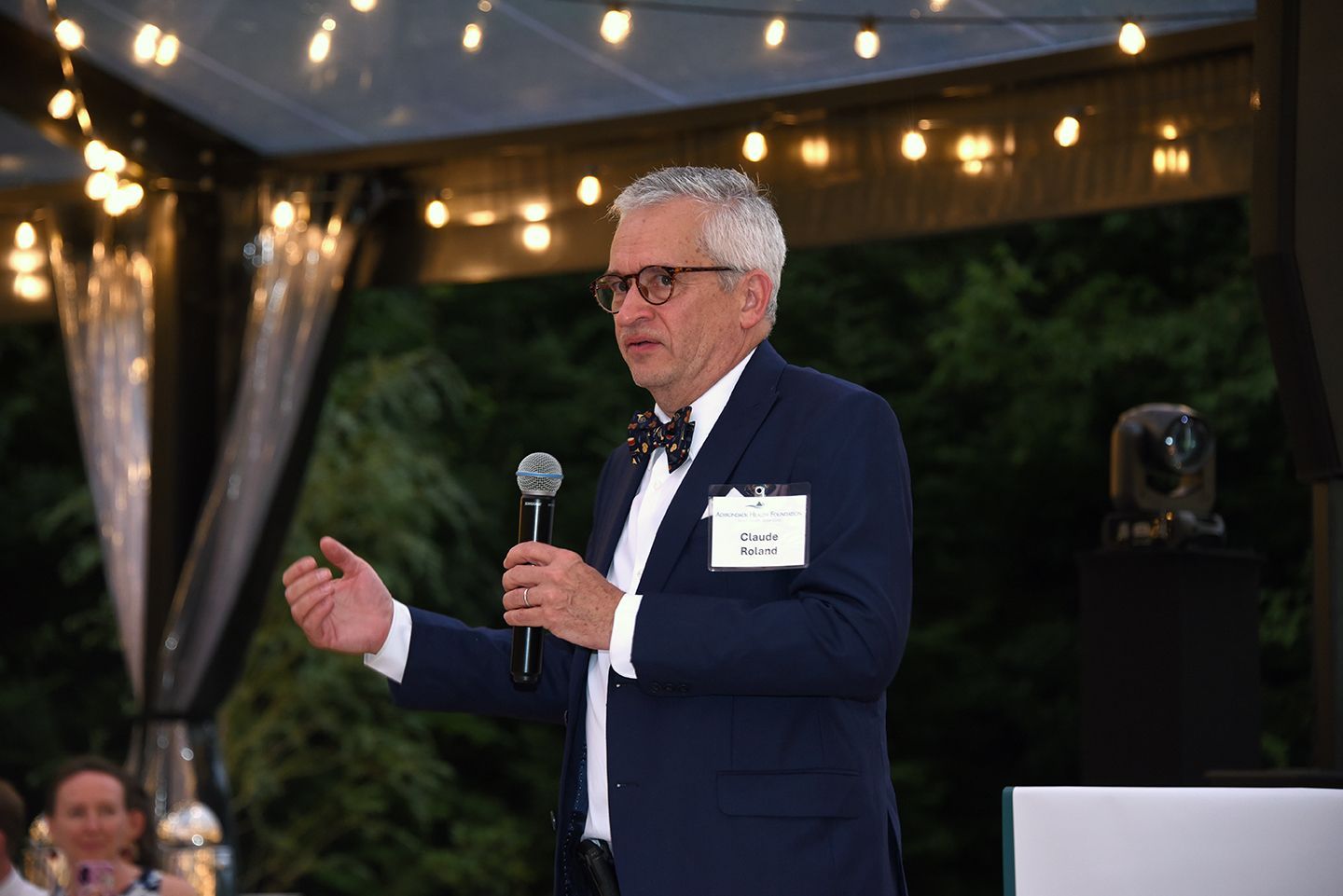 Man in a suit speaks into a microphone at an outdoor event. He has grey hair, glasses, and a bow tie.