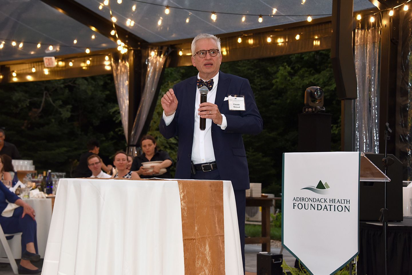Man in a suit speaking at a podium, likely at an outdoor event under a tent.