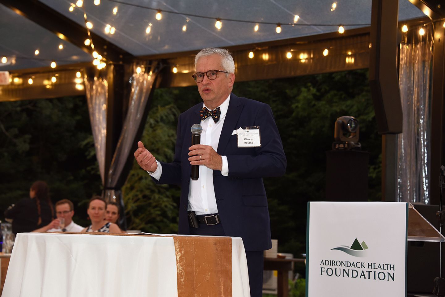 Man in a suit and bow tie speaking at an outdoor event, holding a microphone, with a sign for Adirondack Health Foundation.