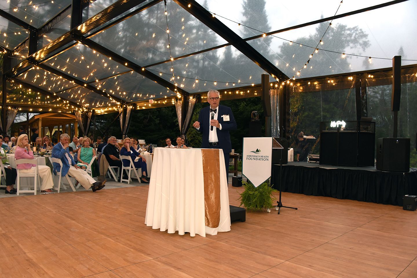 Man speaking at a podium under a clear tent with string lights, addressing an audience seated at tables outdoors.