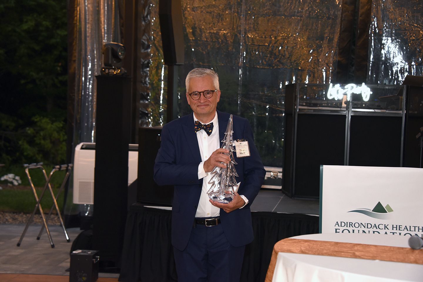 A man in a suit holds a silver saxophone on a stage with a banner for Adirondack Health.
