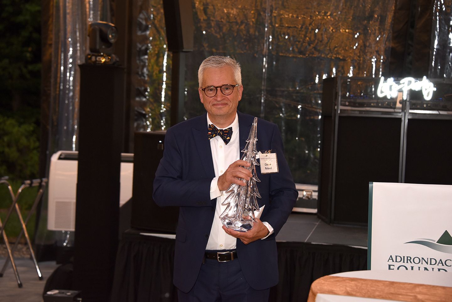 Man in a suit and bow tie holds a trophy, smiling on a stage with a foundation banner.