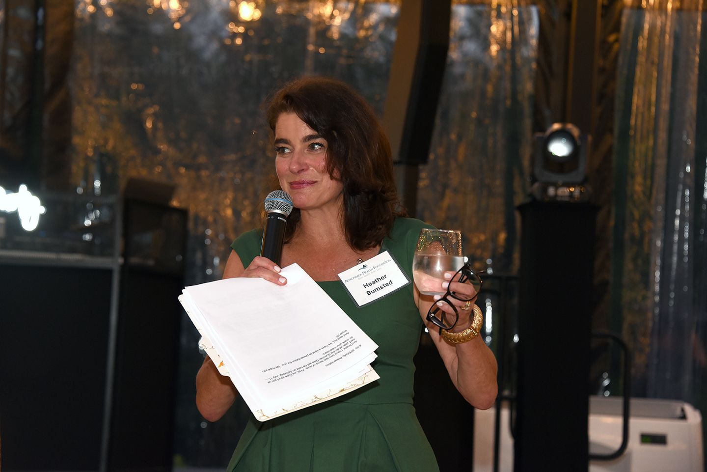 A woman in a green dress speaks at an event, holding papers and a glass of wine. She wears a name tag and smiles slightly.