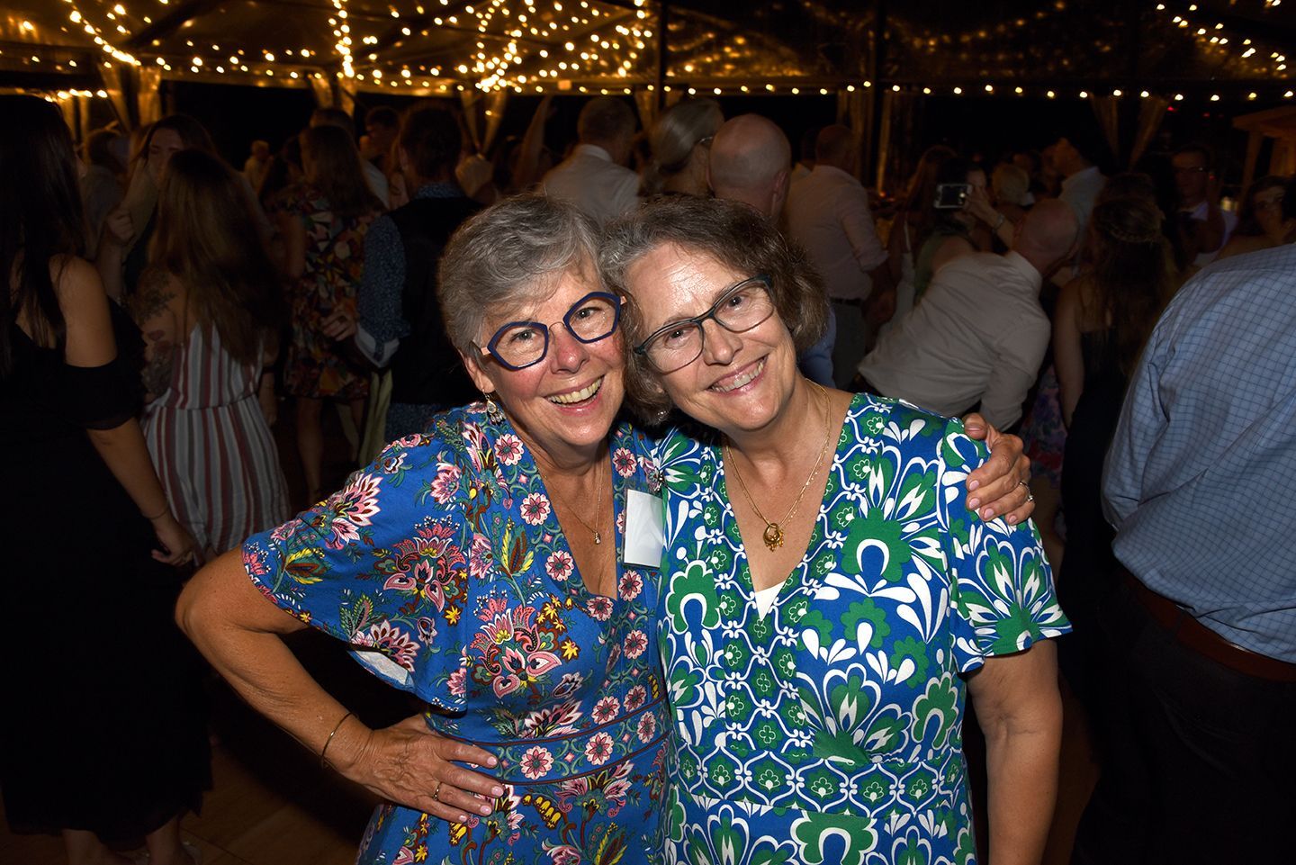Two women smiling, embracing at a party with string lights overhead