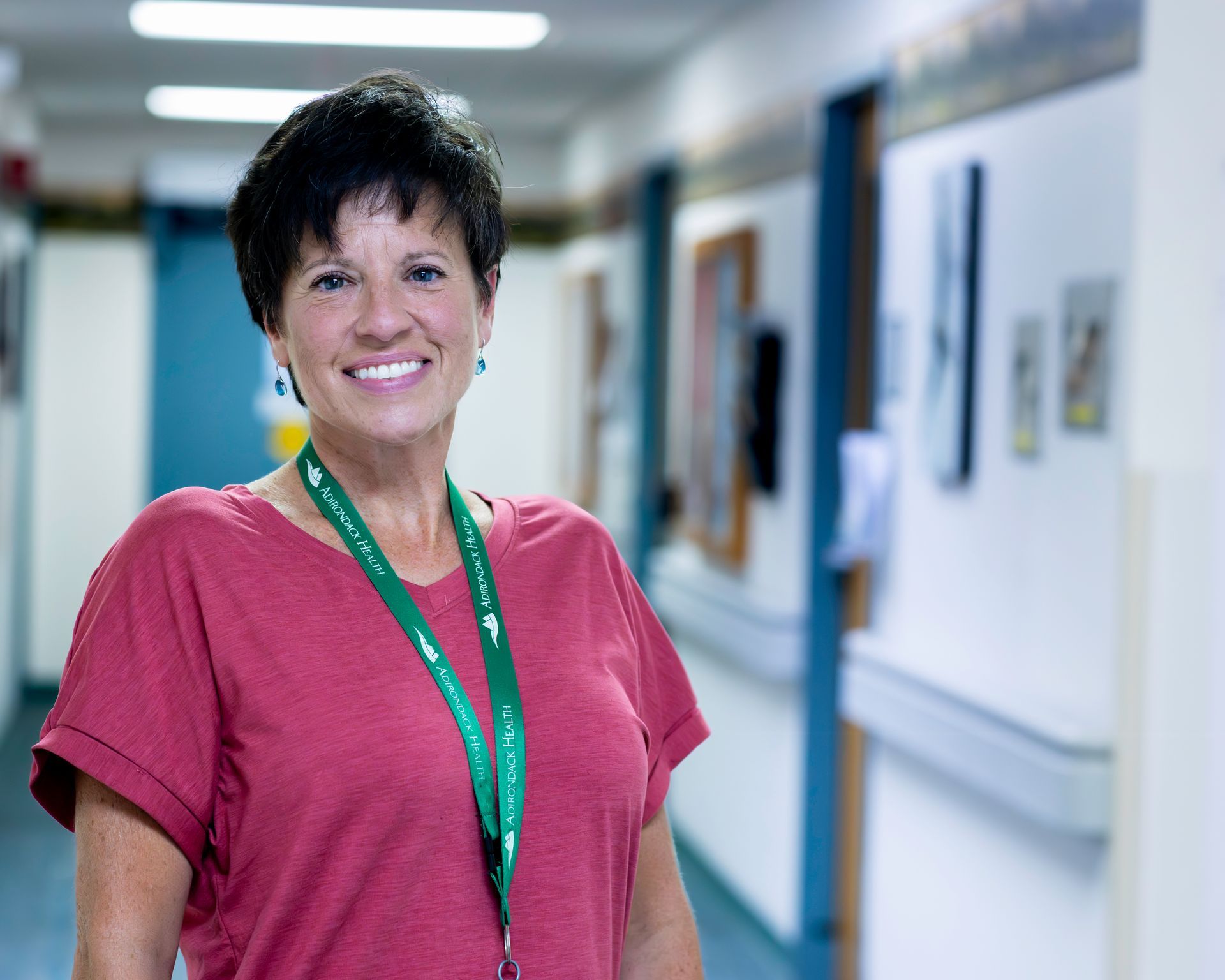 Woman in red shirt smiles in a hospital hallway, wearing a green lanyard.