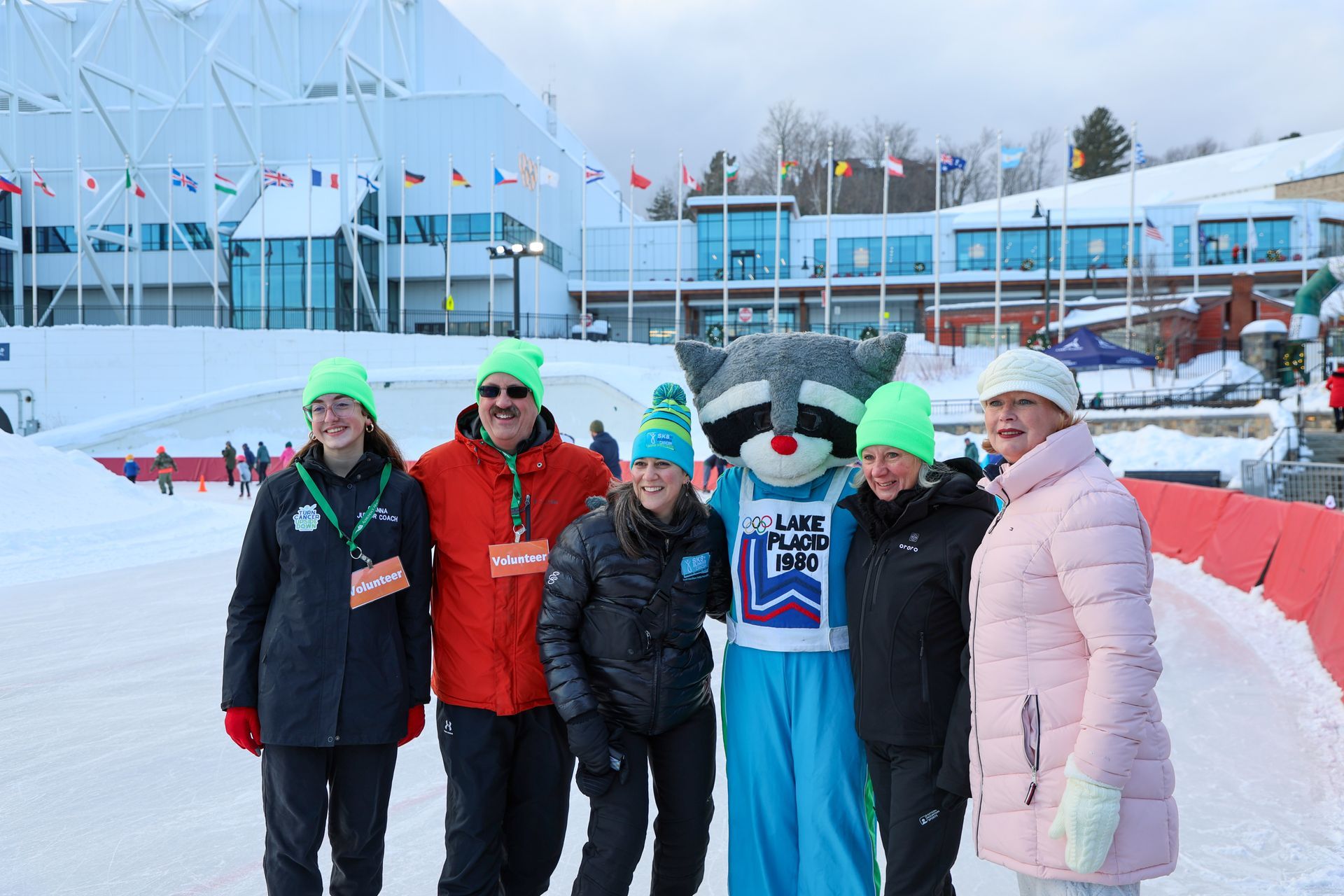 Group poses with a raccoon mascot wearing a hat and bib in front of a snowy sports complex.