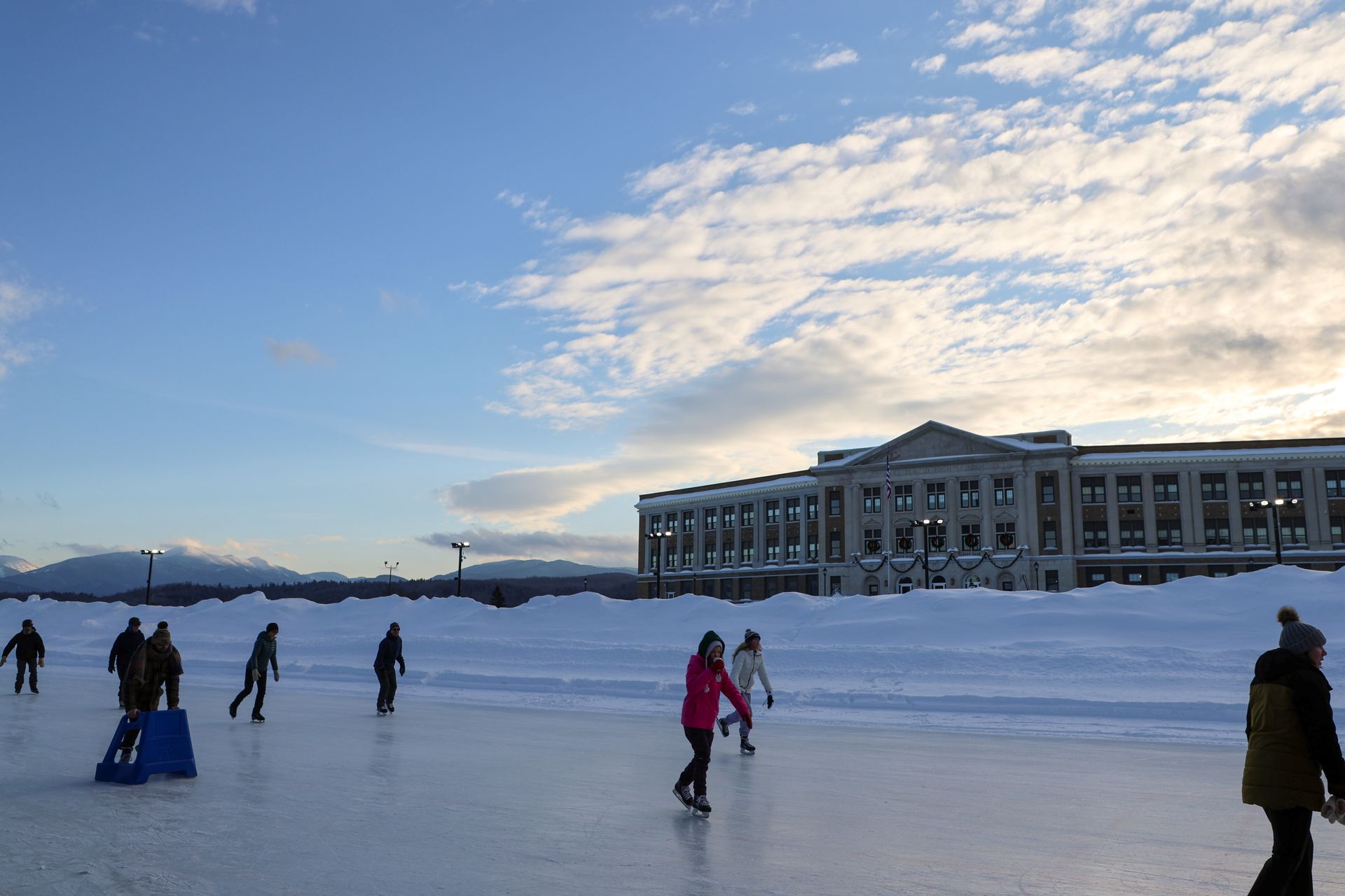 People ice skate on a frozen body of water in front of a large building under a blue sky with clouds.