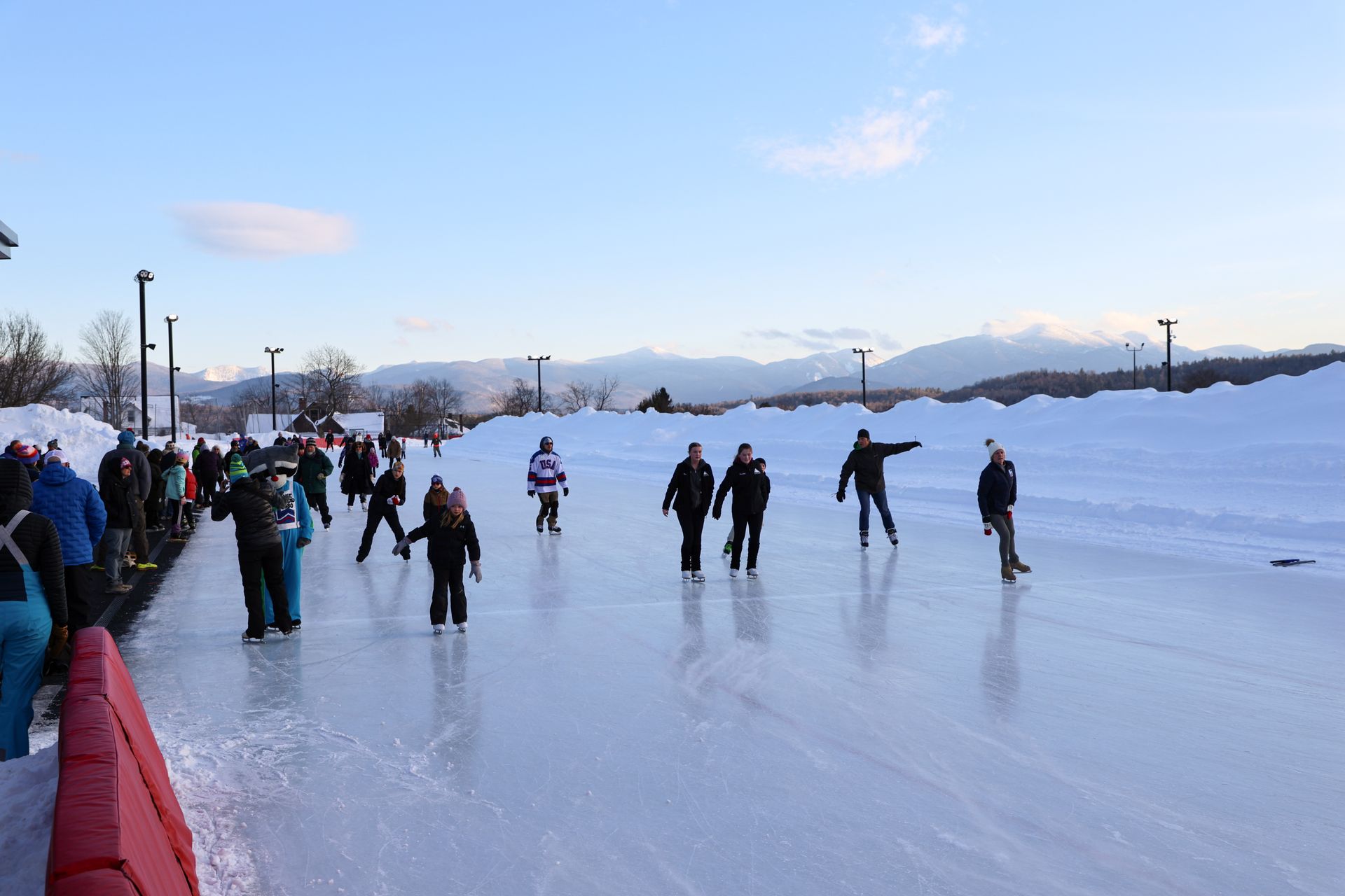 People ice skating on a large outdoor rink with a snow bank to the right and mountains in the distance under a blue sky.