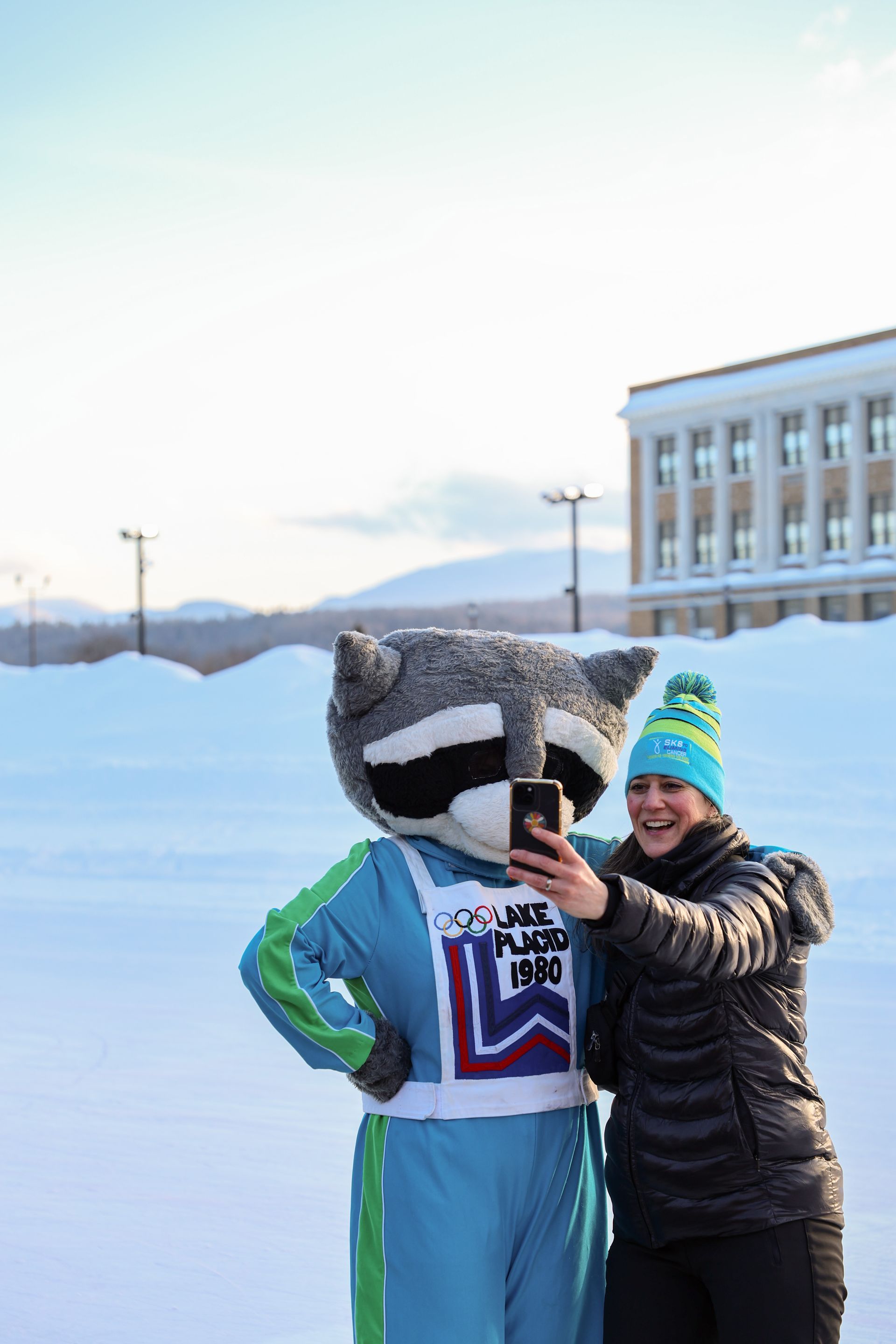 Woman taking selfie with raccoon mascot in snowy setting, wearing winter gear. Building in background.