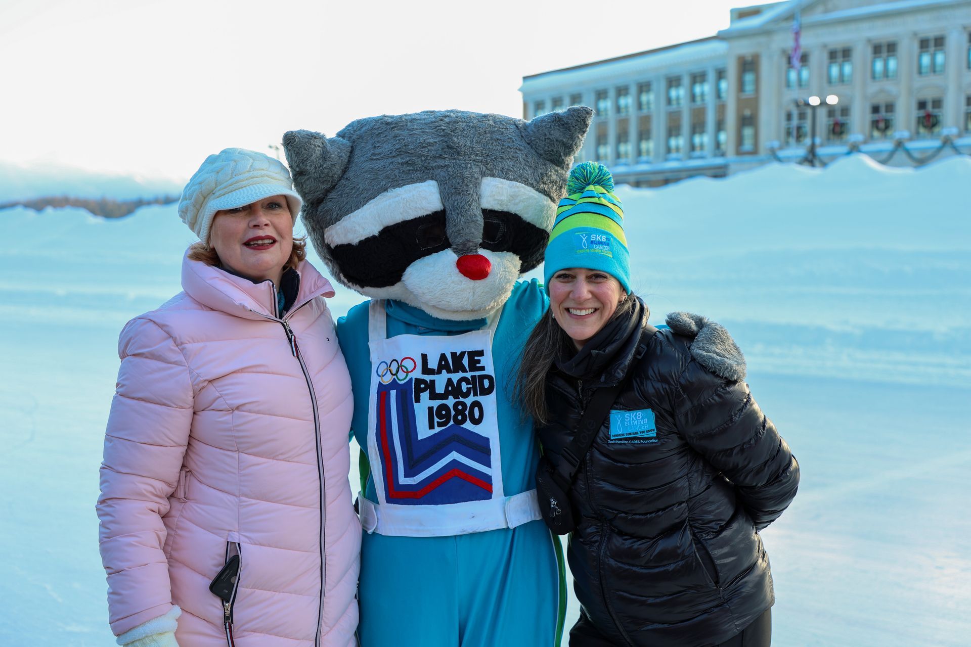 Two women pose with a raccoon mascot wearing a Lake Placid 1980 Olympic bib. They are in a snowy outdoor setting.