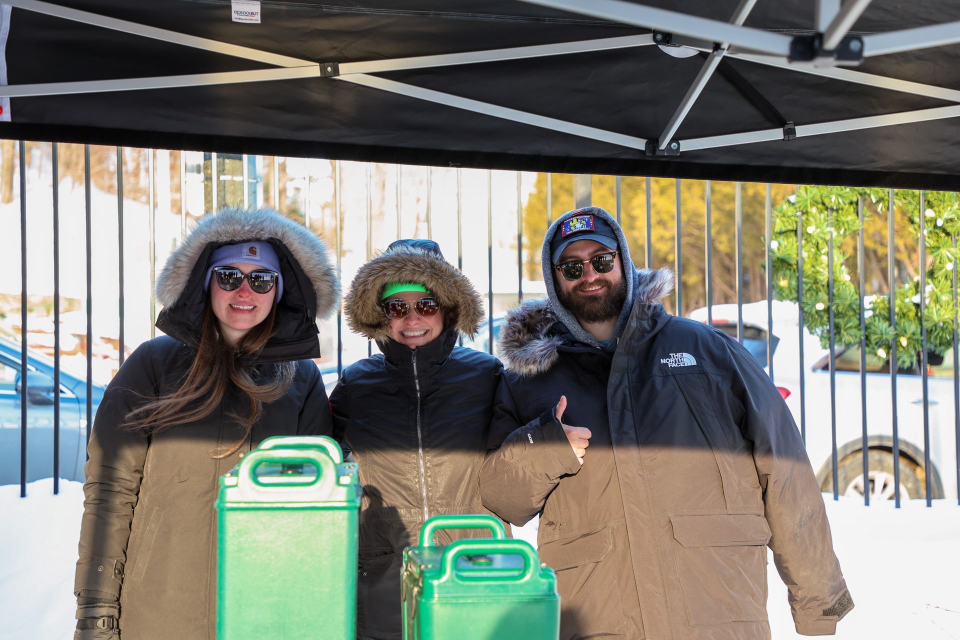 Three people bundled in winter coats, smiling in front of two green containers.