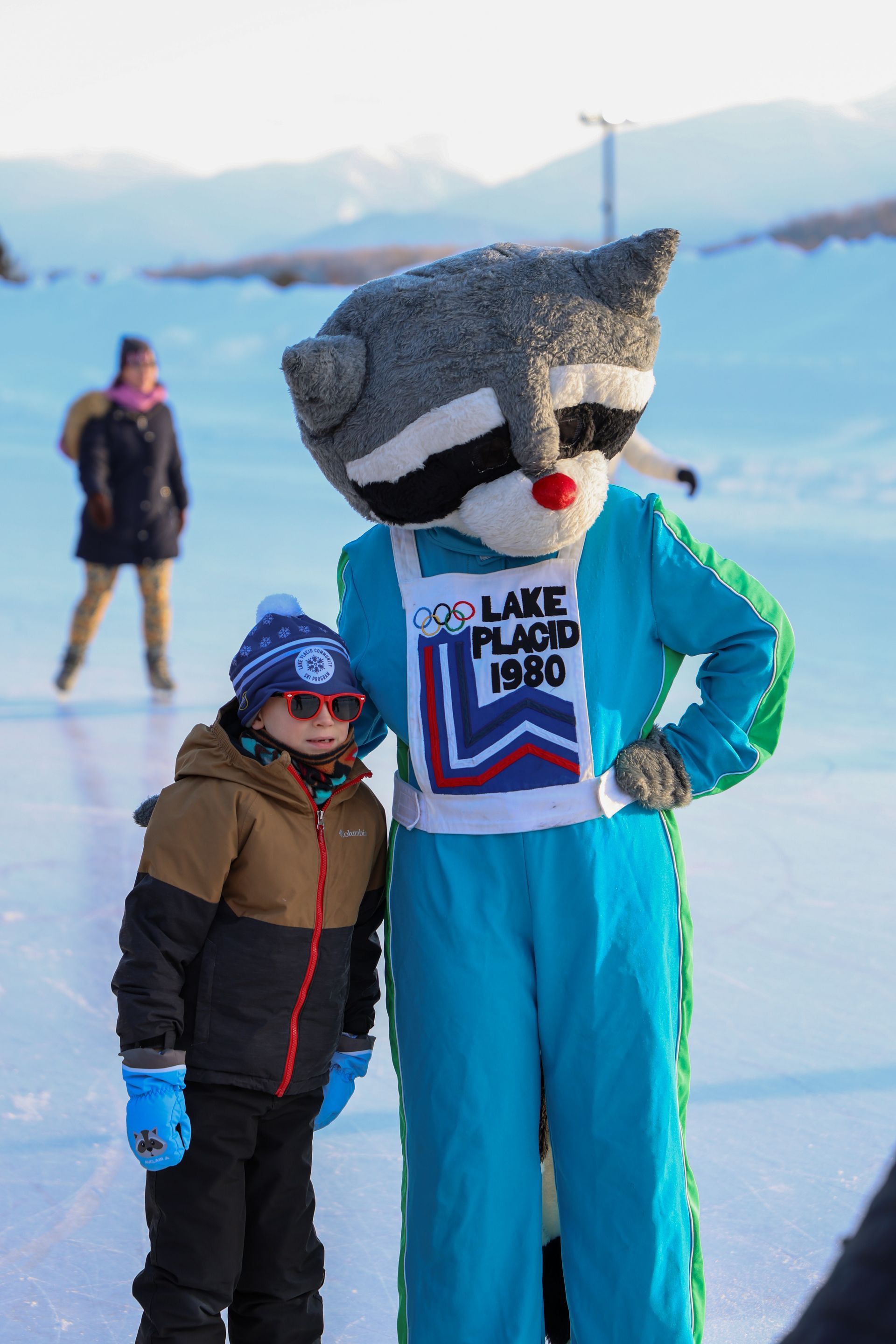 A child and the mascot of the 1980 Lake Placid Olympics, a raccoon in a blue jumpsuit, stand on ice.