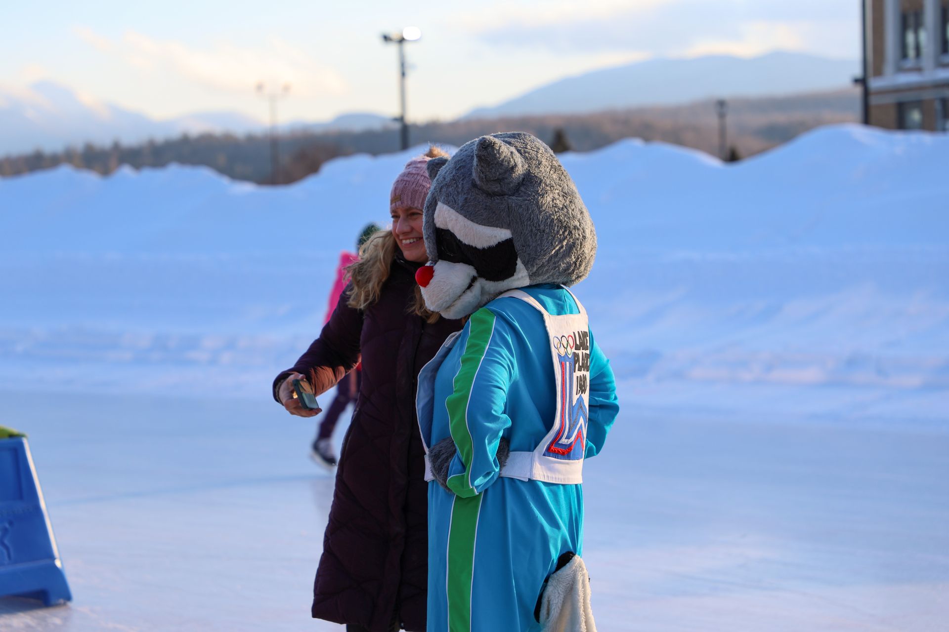 A person in a raccoon mascot costume talks with a woman on an ice skating rink, snowy mountains in the background.