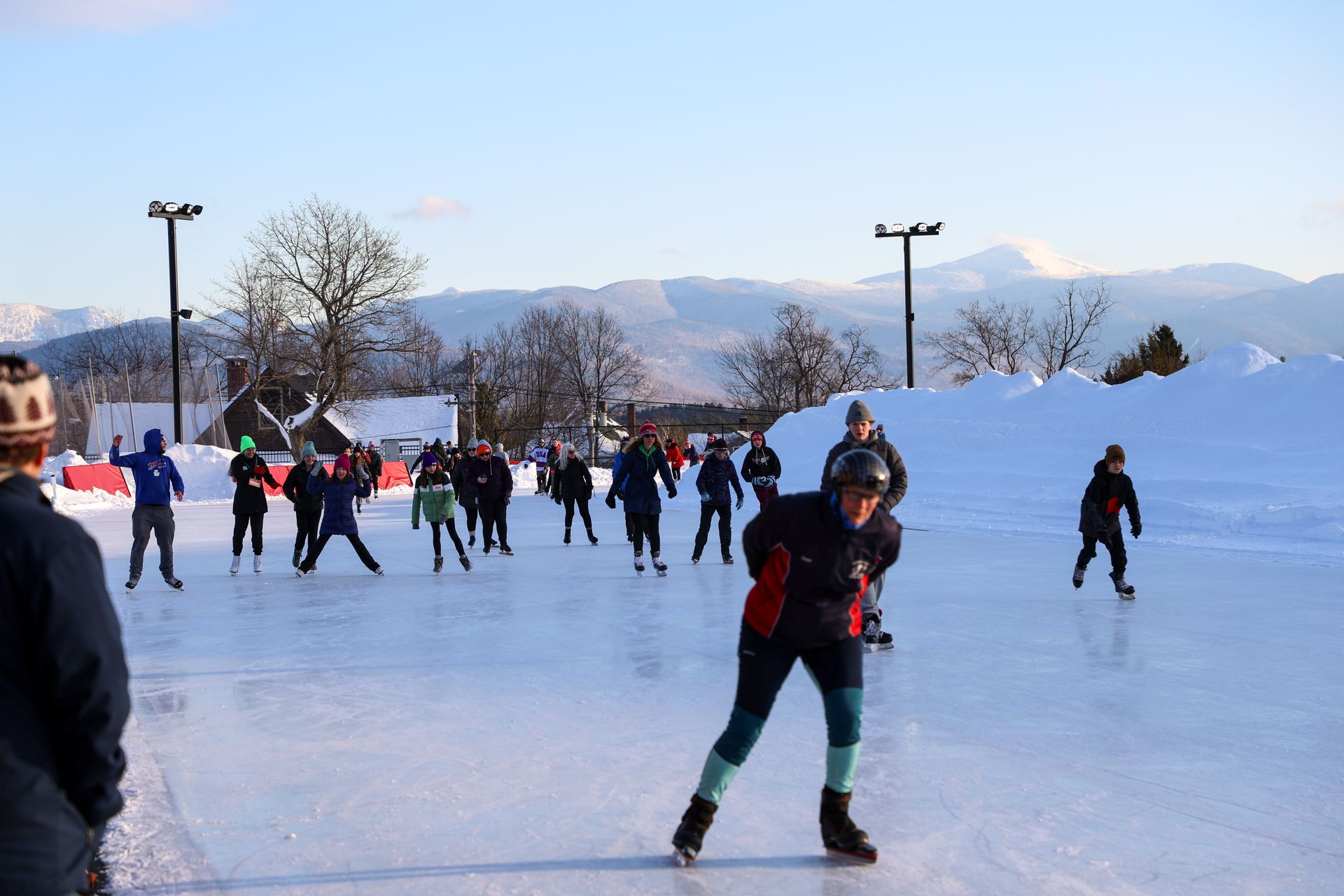 People ice skating on an outdoor rink on a sunny day. Snow-covered mountains are in the background.
