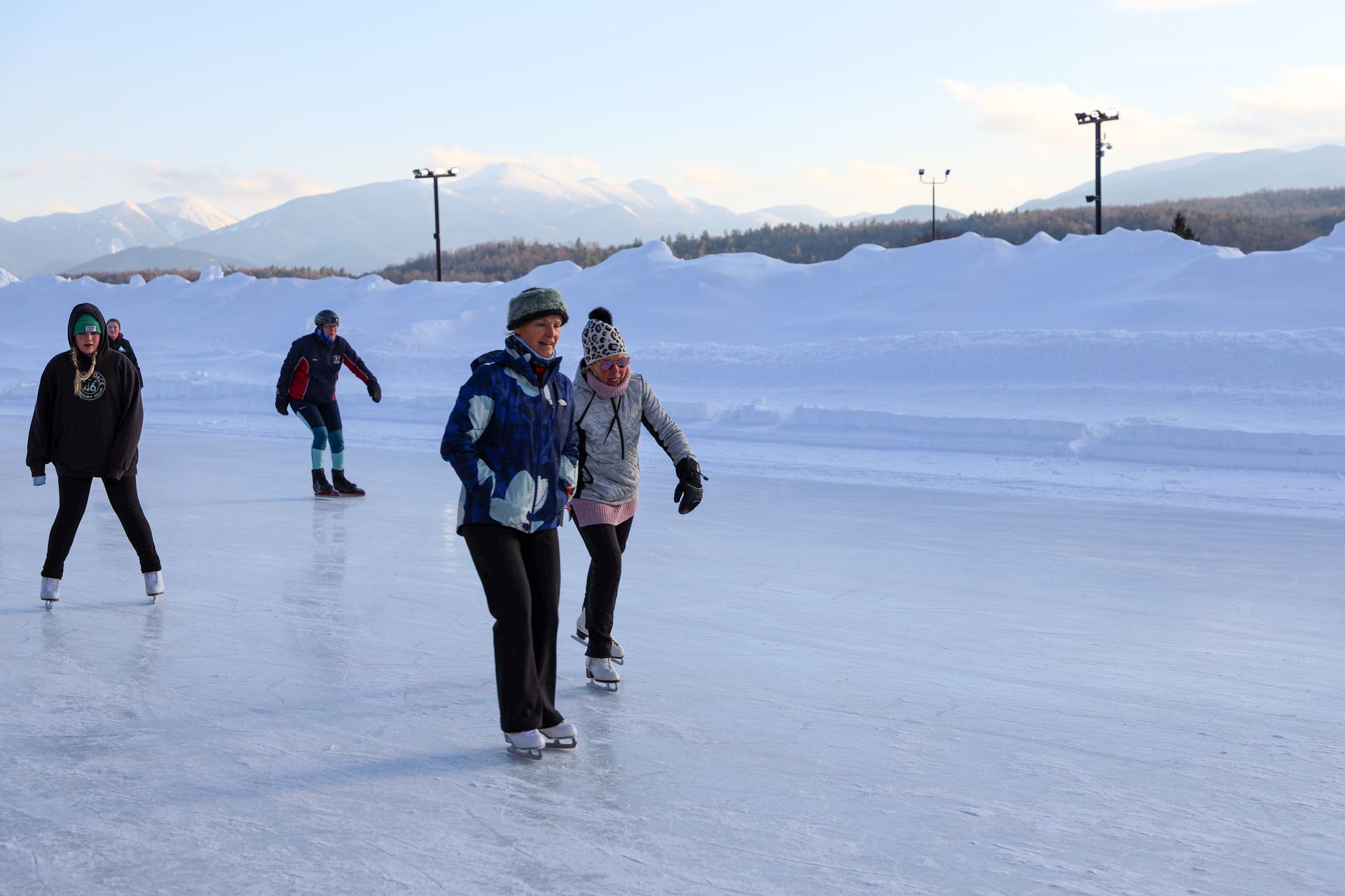 People ice skating on a frozen rink with snow banks and mountains in the background.