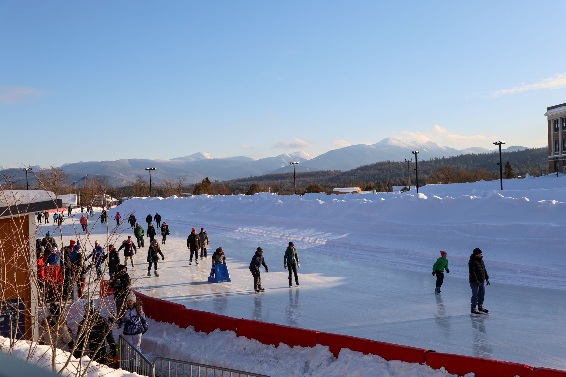 People ice skate on an outdoor rink on a sunny day, with snow-covered ground and mountains in the background.