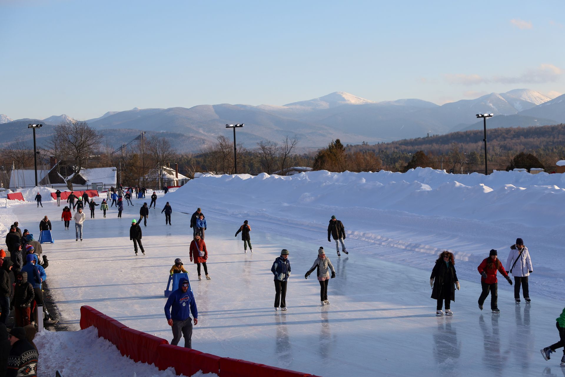 People ice skating on a large outdoor rink, with snow-covered banks and mountains in the background under a bright blue sky.