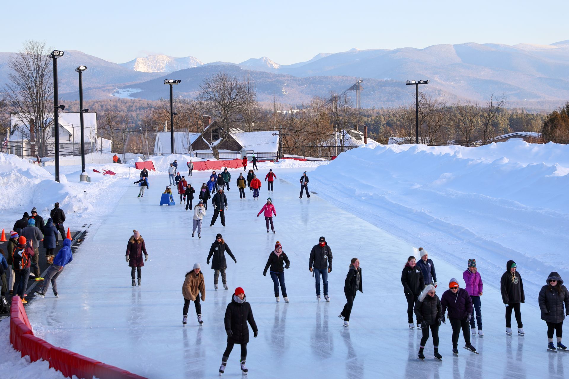 People ice skate on a long, cleared path through a snowy landscape, with mountains in the background.