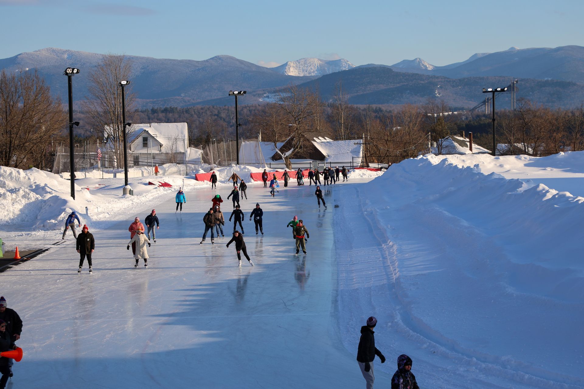 People ice skate on an outdoor rink with snow-covered banks, buildings, and mountains in the background on a sunny day.