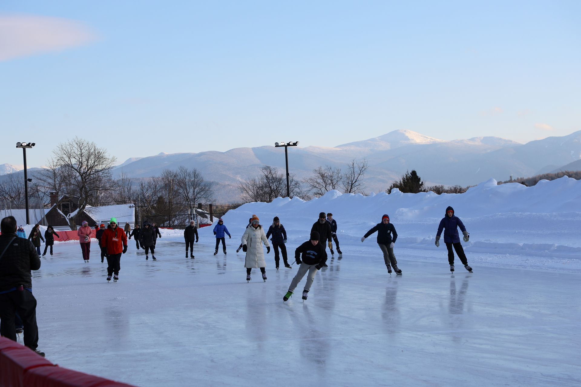 People ice skating on a frozen rink outdoors with snow-covered mountains in the background under a blue sky.