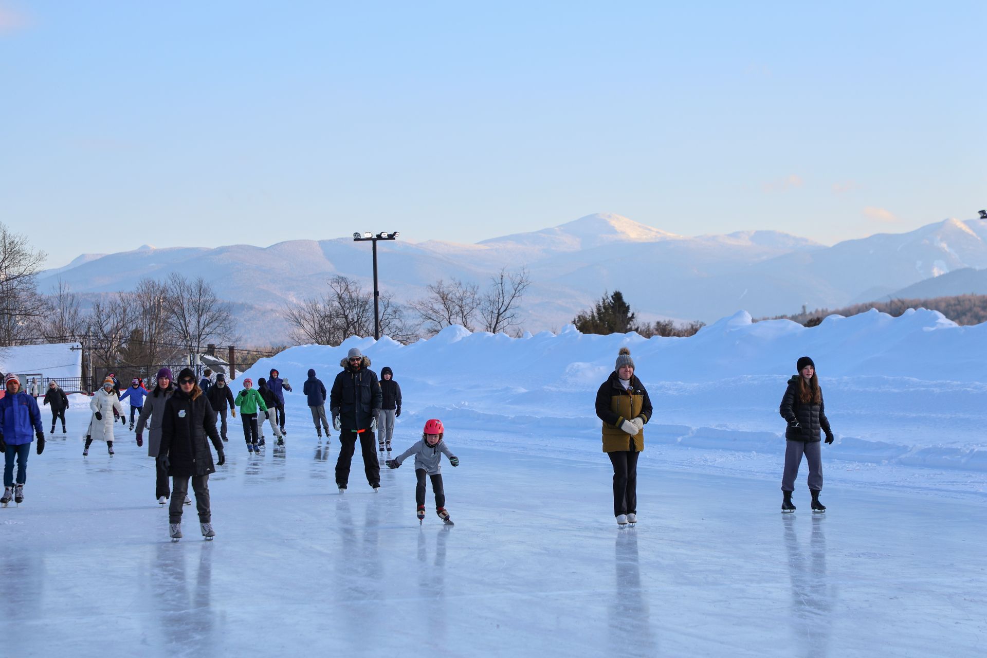 People ice skating on an outdoor rink with snow banks and mountains in the background on a sunny day.