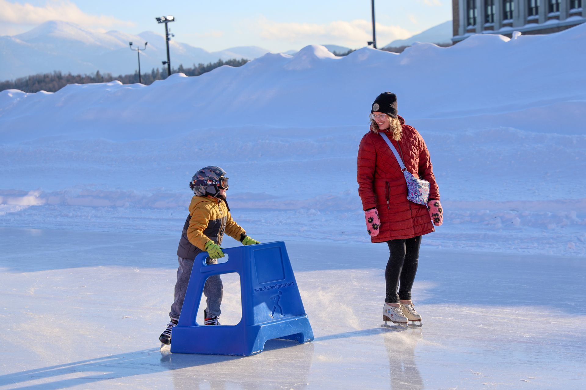 A child learning to ice skate with a blue support, watched by a woman in a red coat, on a sunny outdoor rink.