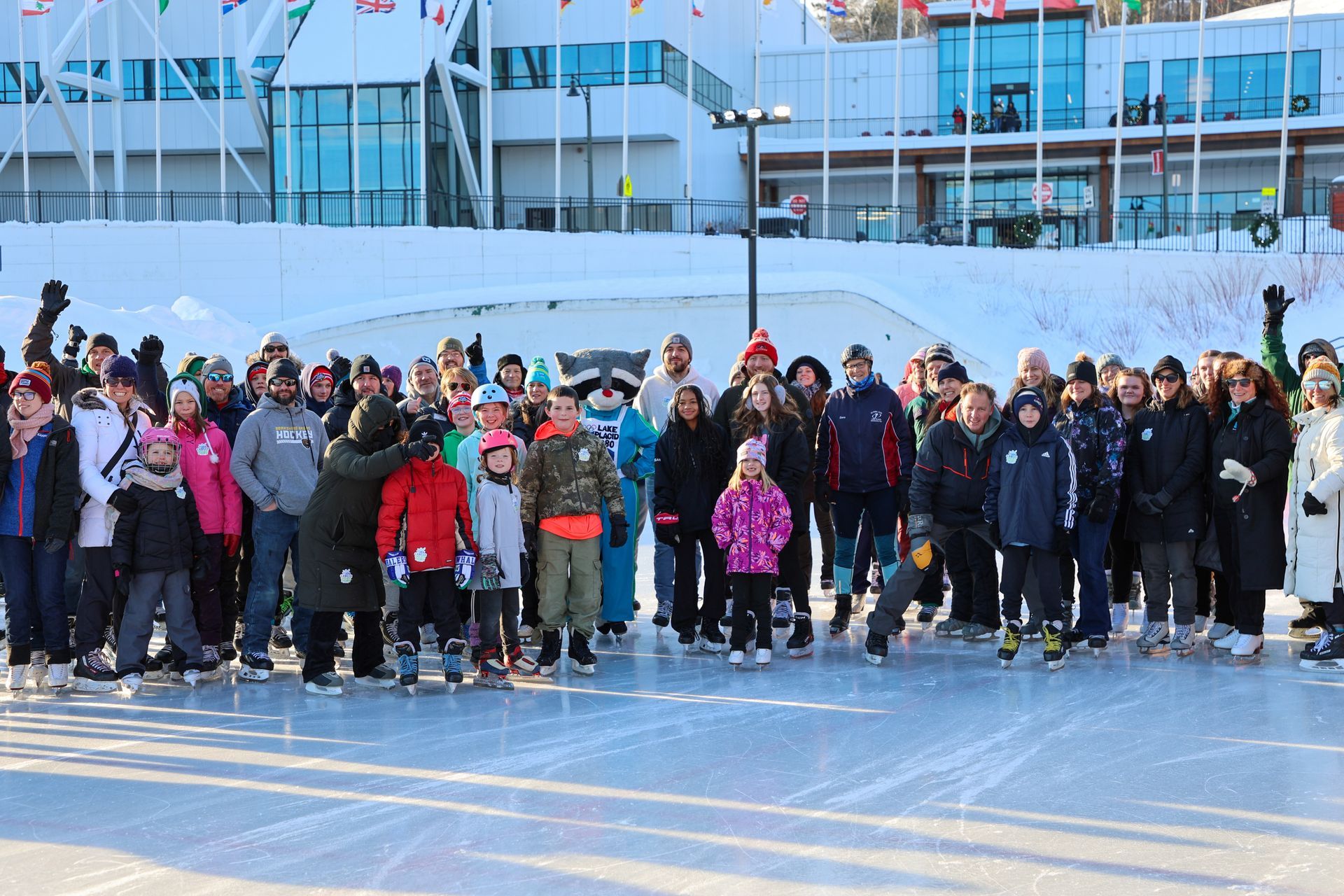 A large group of people on ice skates pose for a photo in front of a building with flags and snow.