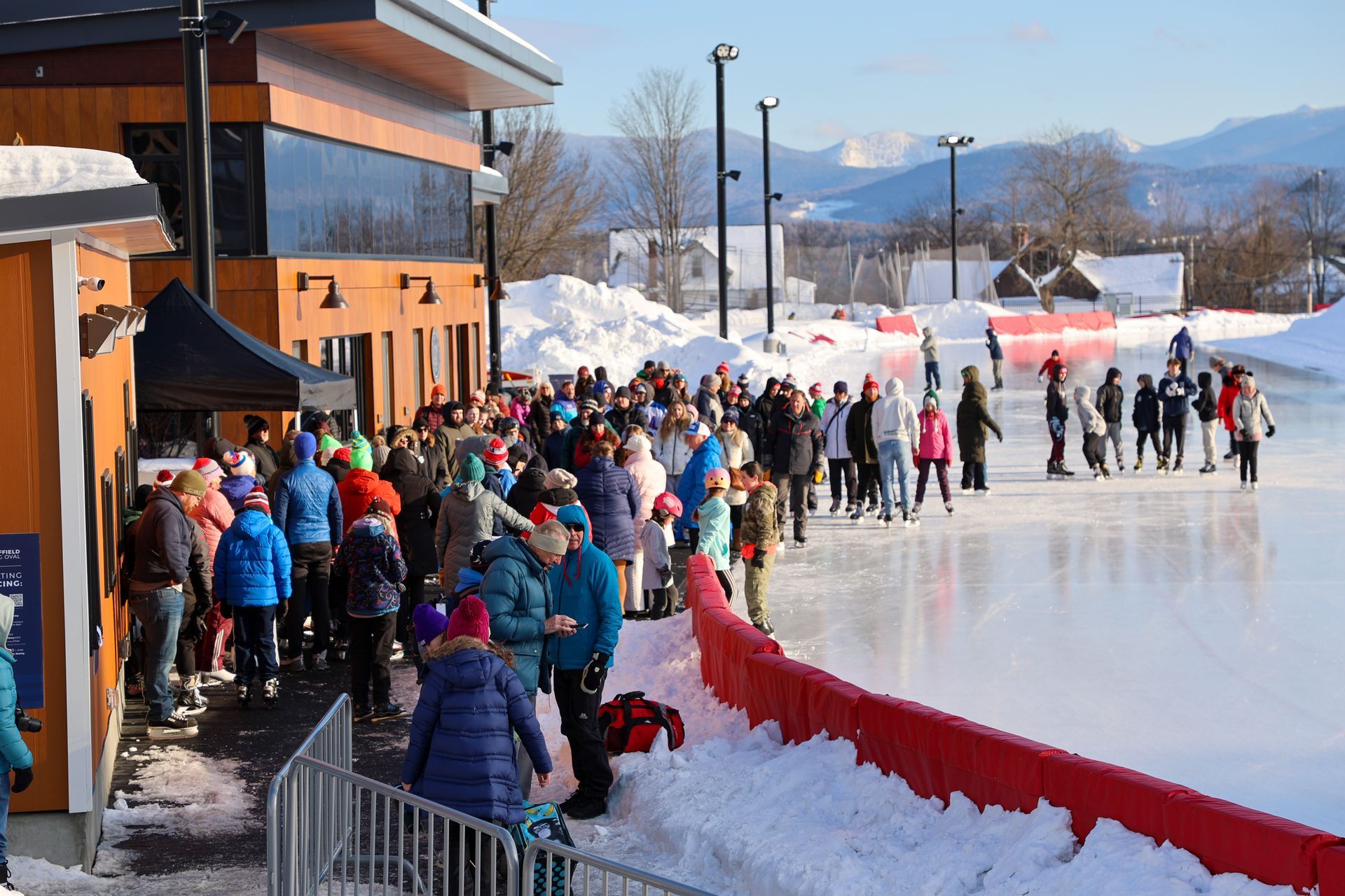 People ice skating on an outdoor rink, with a crowd near a building and snowy mountains in the background on a sunny day.