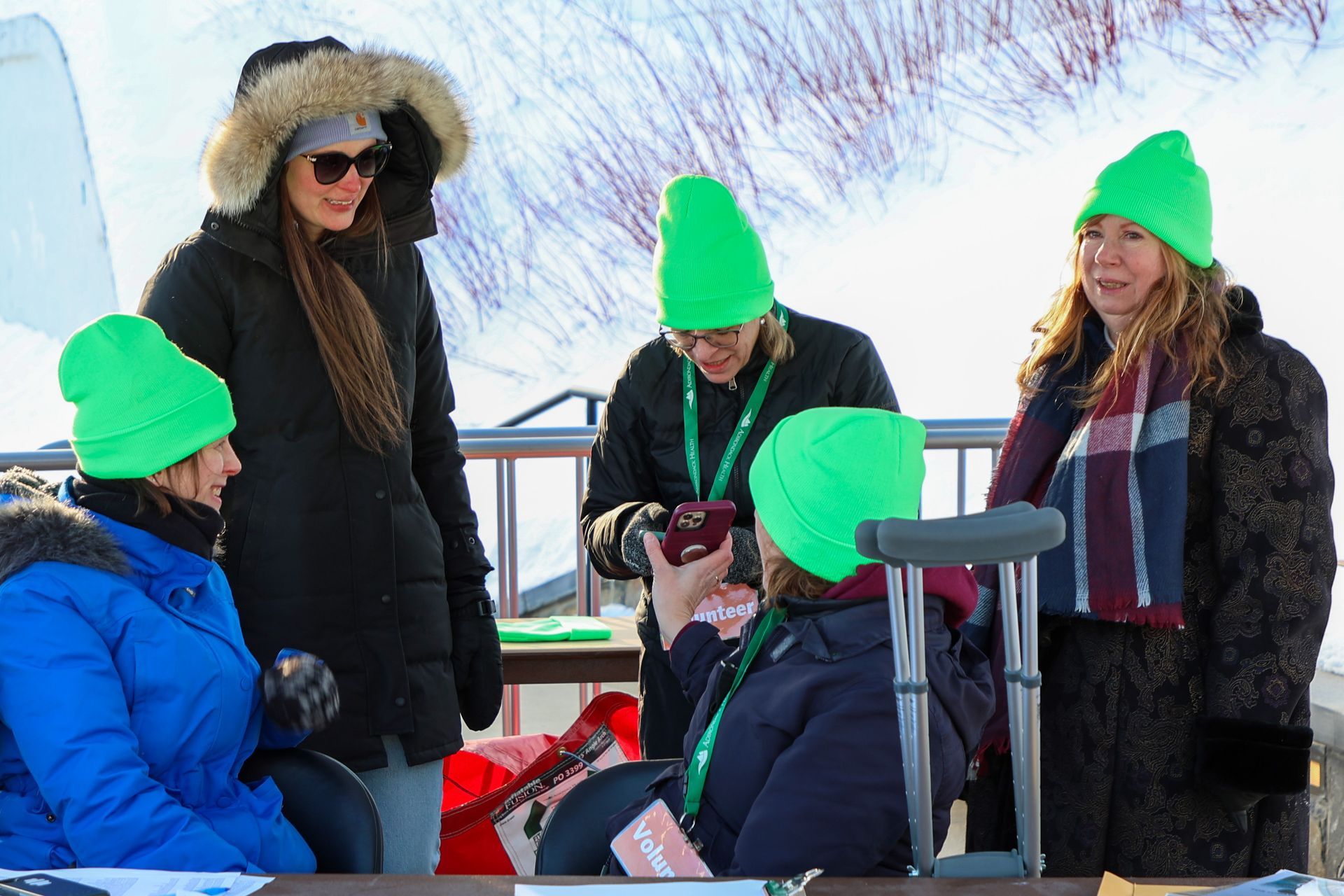 Five people in winter attire, some wearing bright green hats, gather outside near snow. One uses crutches.