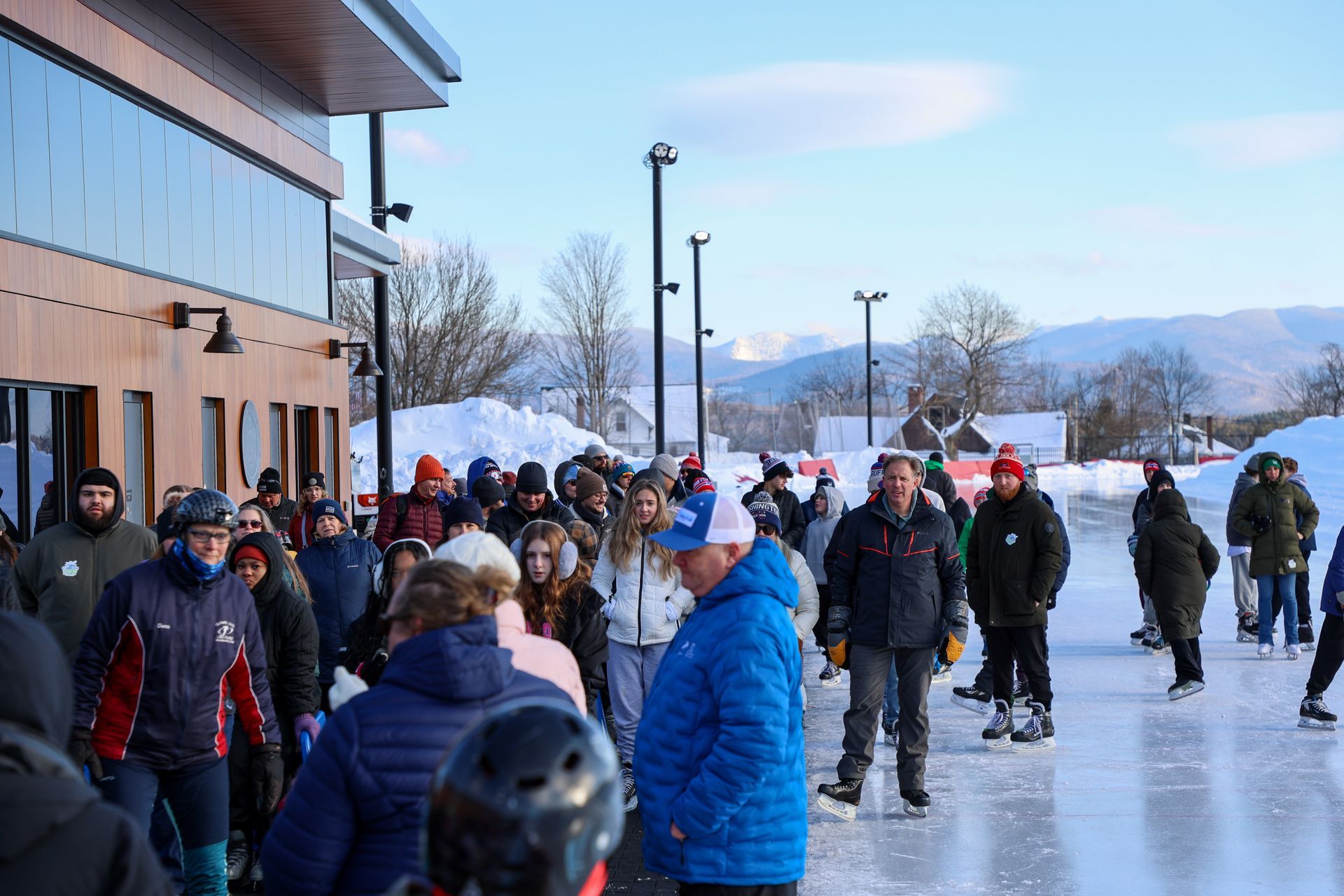 People ice skating outdoors near a building, with snow-covered mountains in the background. Many are wearing winter clothing.