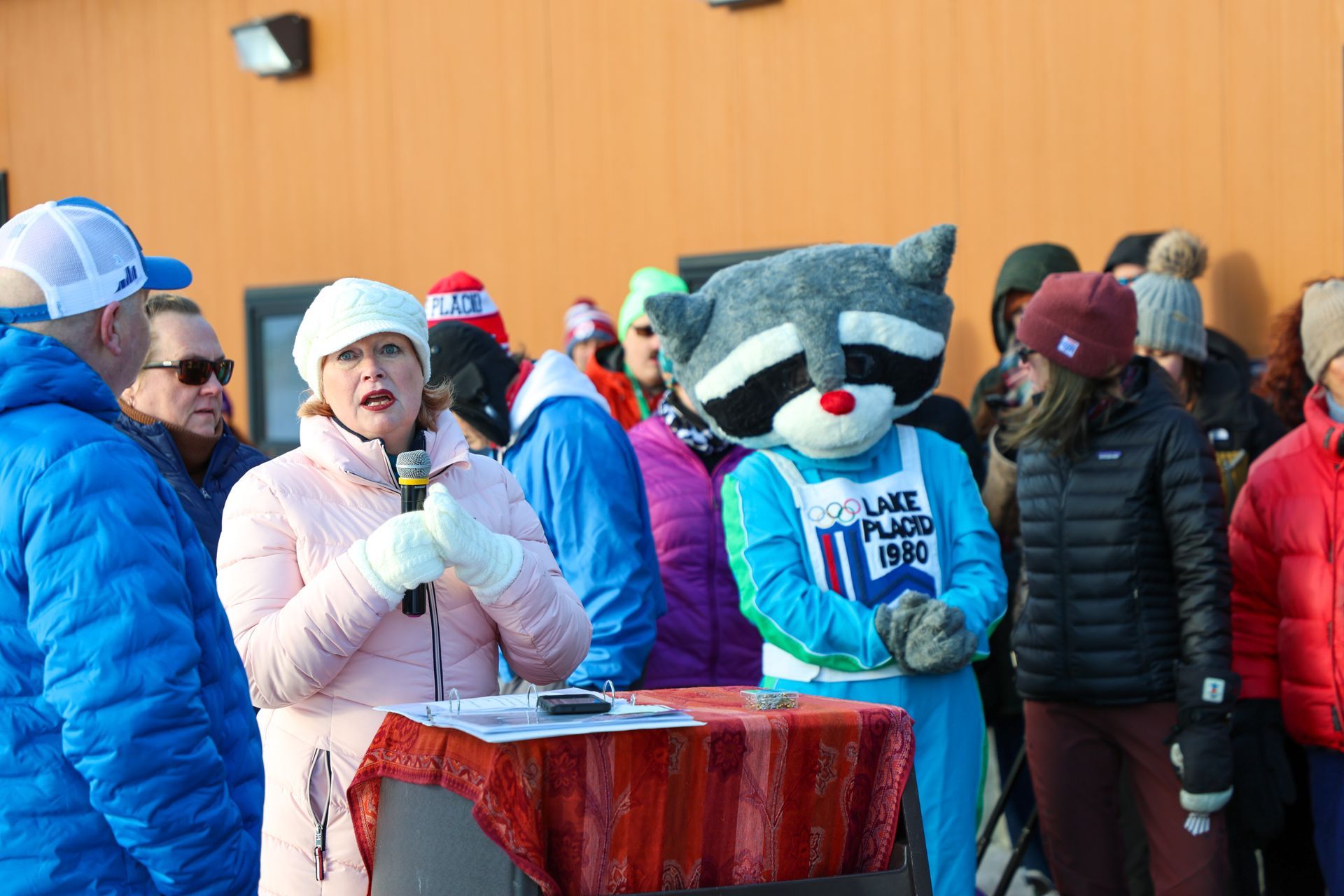 A woman in a pink jacket speaks at a podium next to a raccoon mascot wearing a Lake Placid '80 shirt.