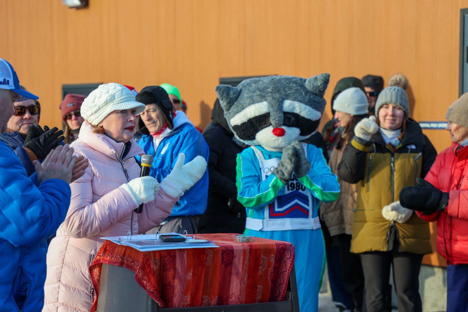 A woman speaks at a podium, clapping as a raccoon mascot stands beside her. People watch in an outdoor setting.