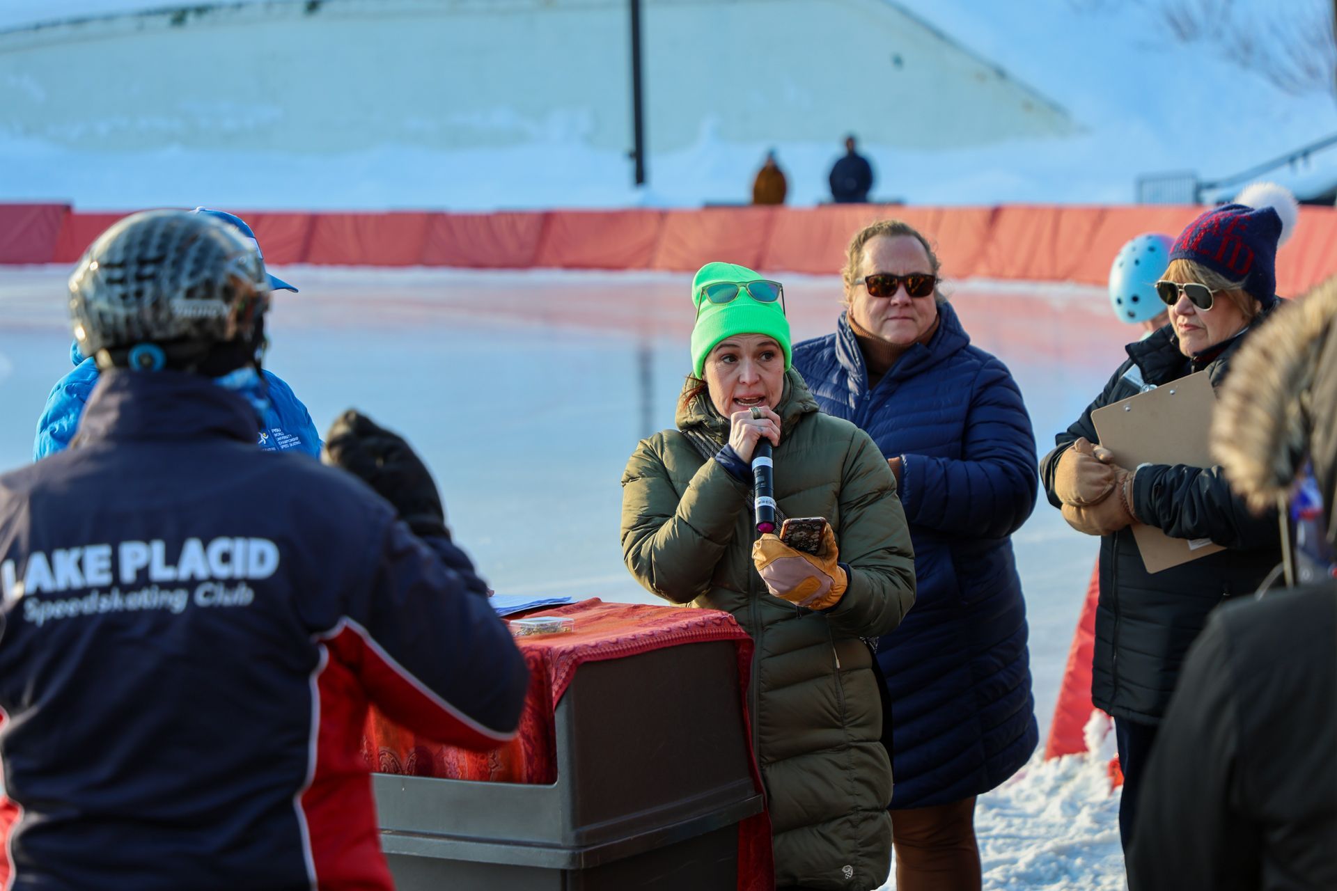 A woman in a green beanie speaks into a microphone at an outdoor ice skating event in Lake Placid, NY.