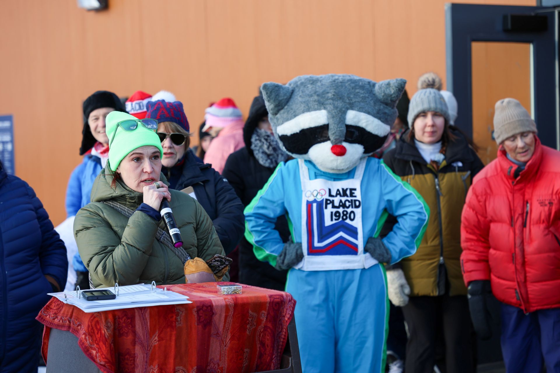 A woman speaks into a microphone at a podium; a raccoon mascot stands beside her, in front of a crowd, outdoors.