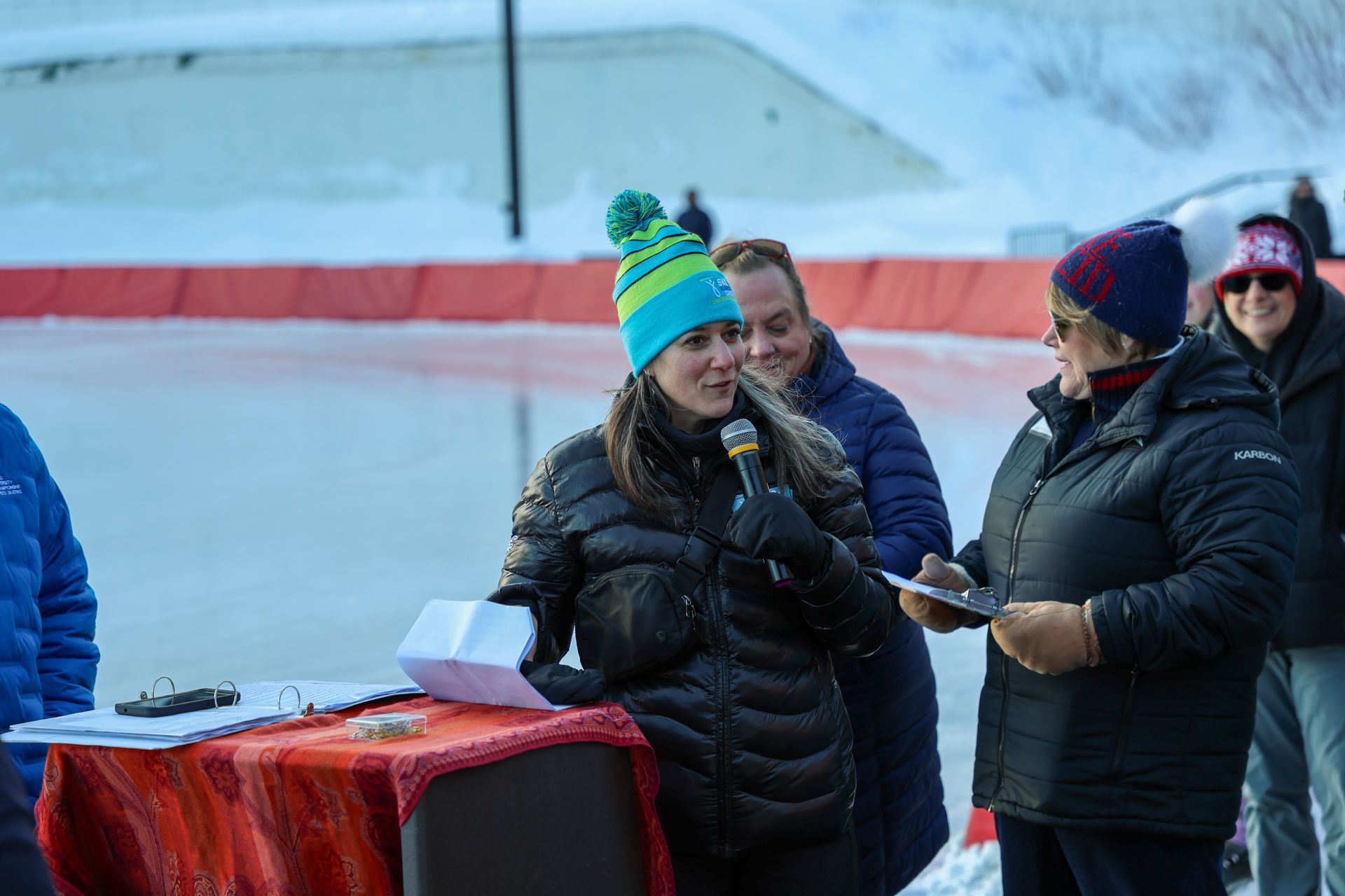 A woman speaks into a microphone at an outdoor event, wearing a colorful hat and black jacket.