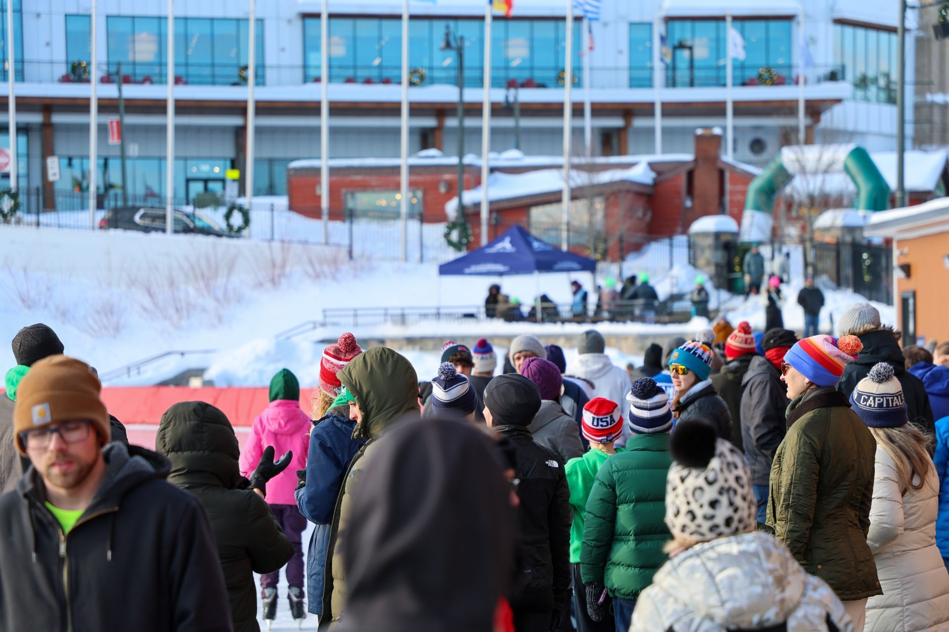 People in winter clothing gather outdoors in front of snow and buildings. Many wear hats.