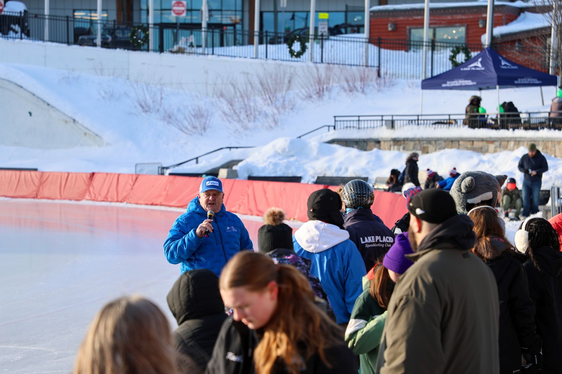 A man in a blue jacket speaks into a microphone on an outdoor ice rink, surrounded by a crowd of people in winter clothing.