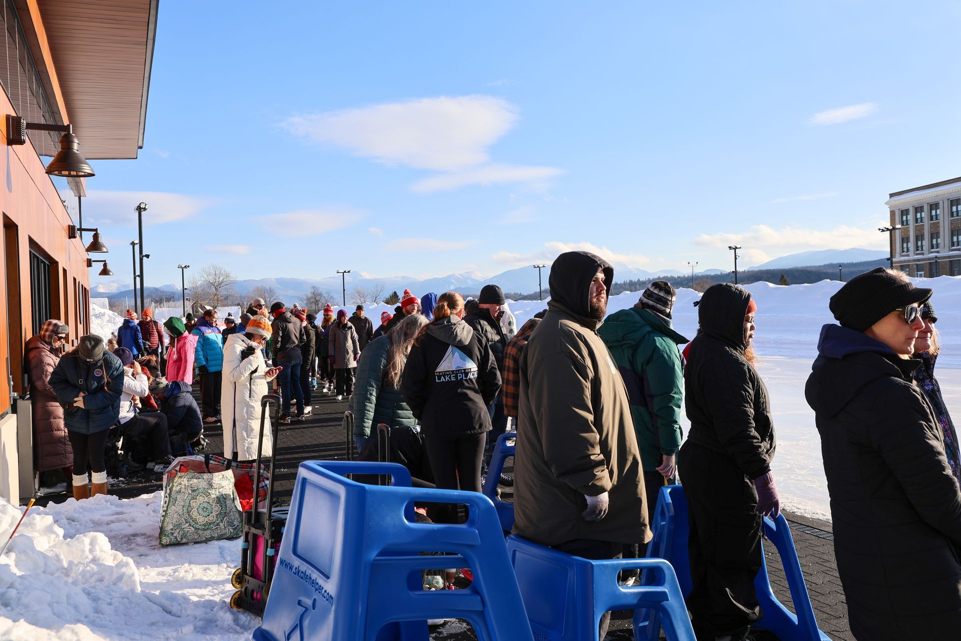 People waiting in line outdoors on a snowy day next to a building. Many wear winter coats and hats.
