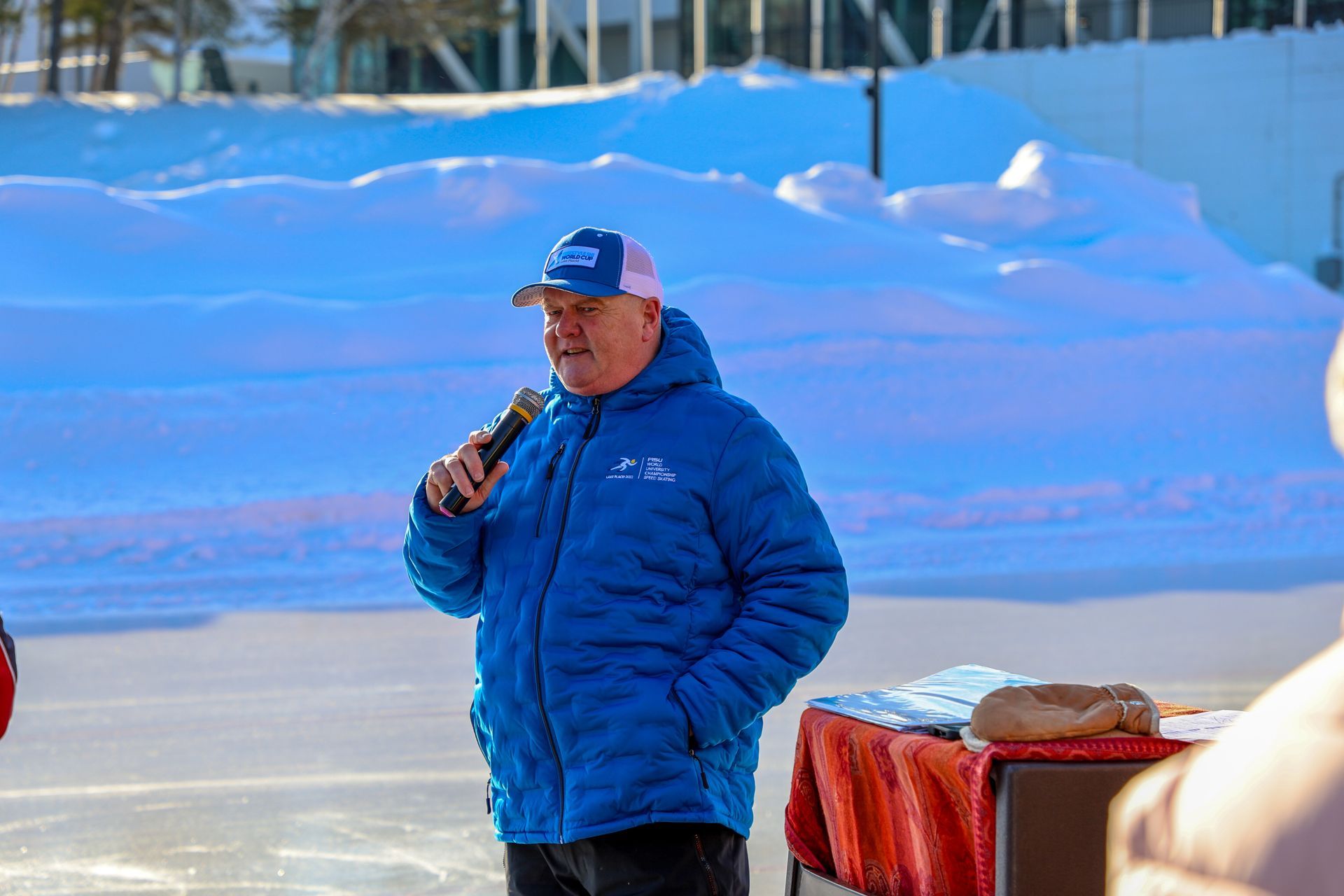Man in blue jacket and hat speaking into a microphone outdoors, snow in the background.