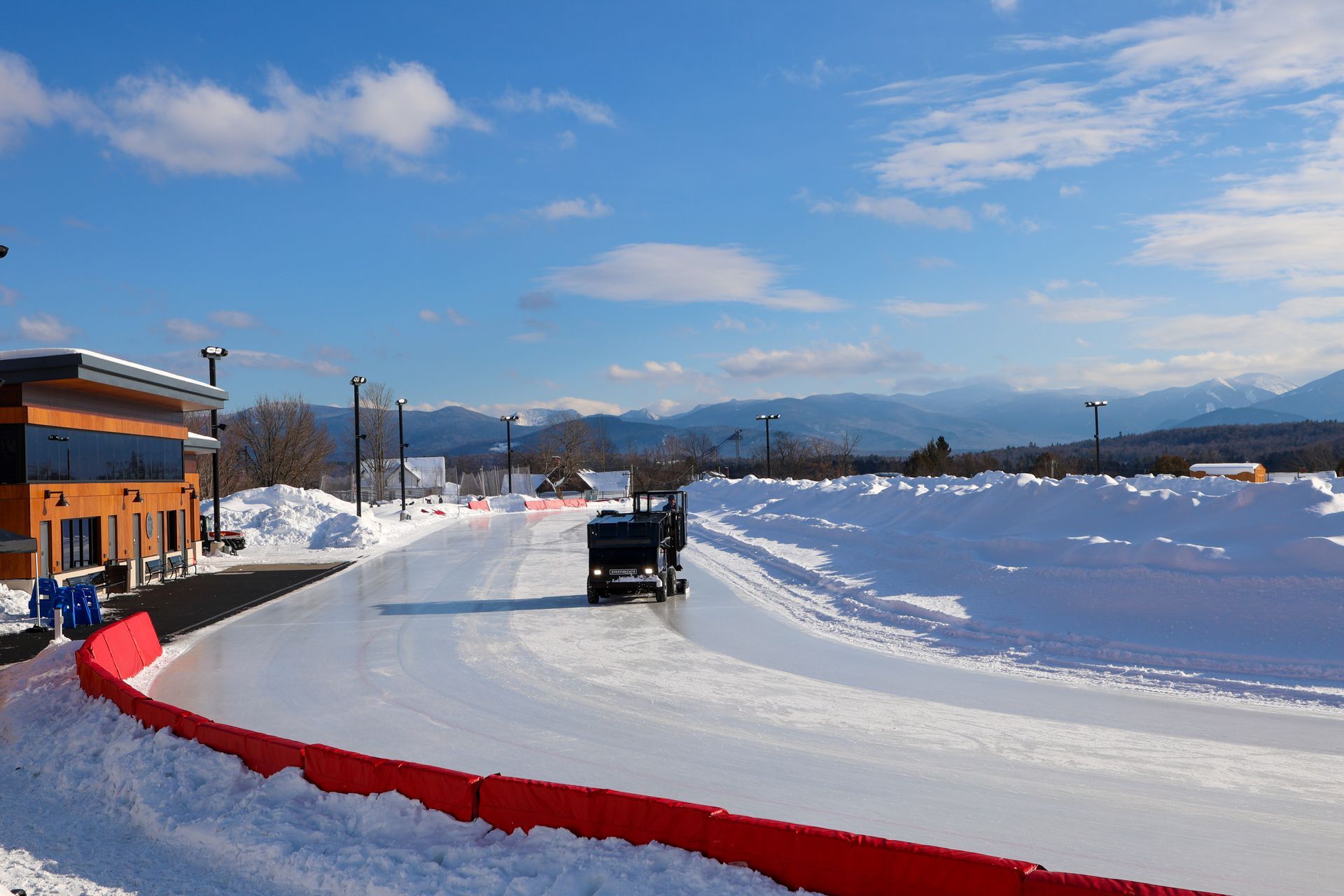 A truck drives on an icy track, bordered by snow and a red barrier, with a building and mountains in the background.