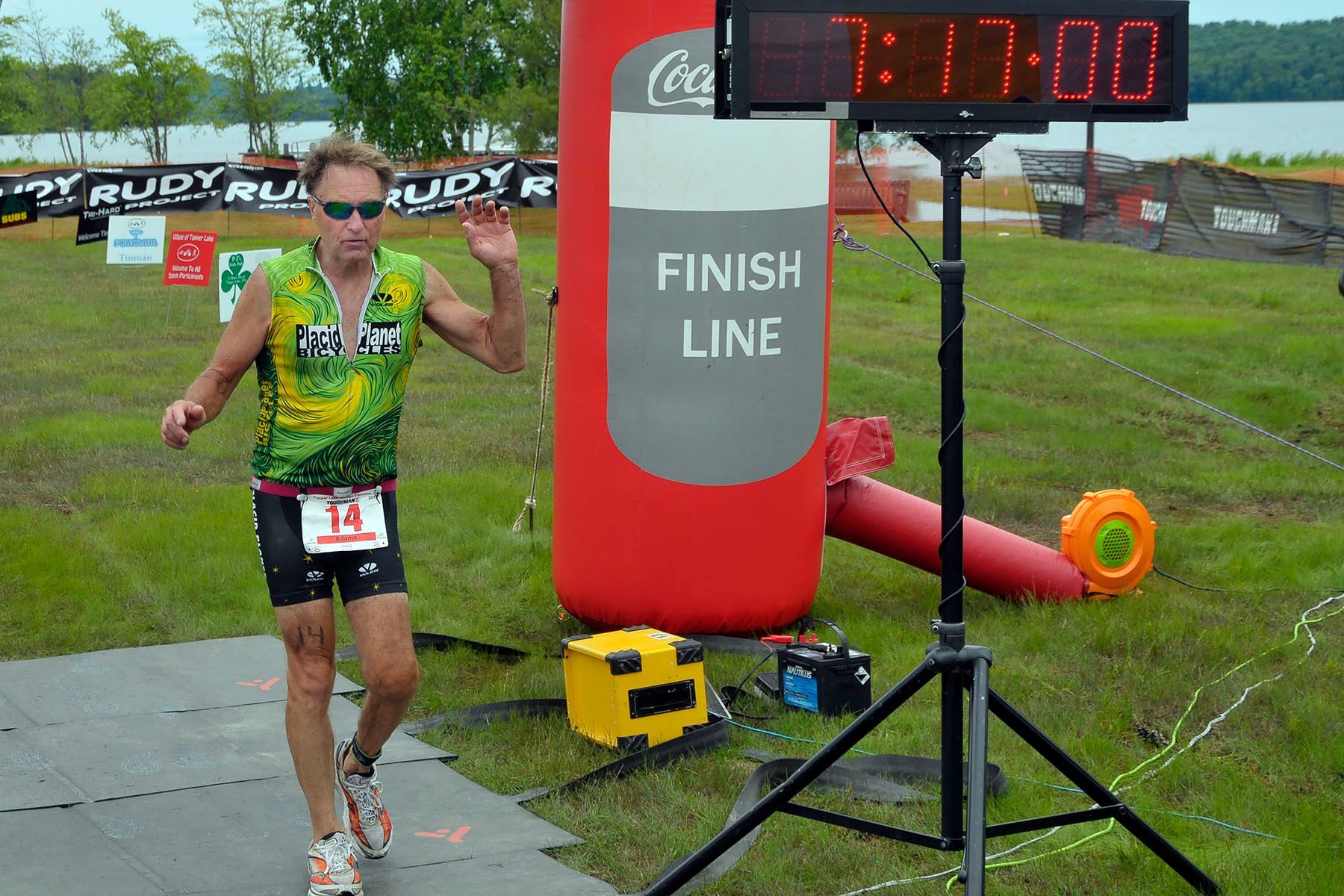 Triathlete crossing the finish line at a race, under a Coke-branded arch, with a digital clock showing 1:17:00.