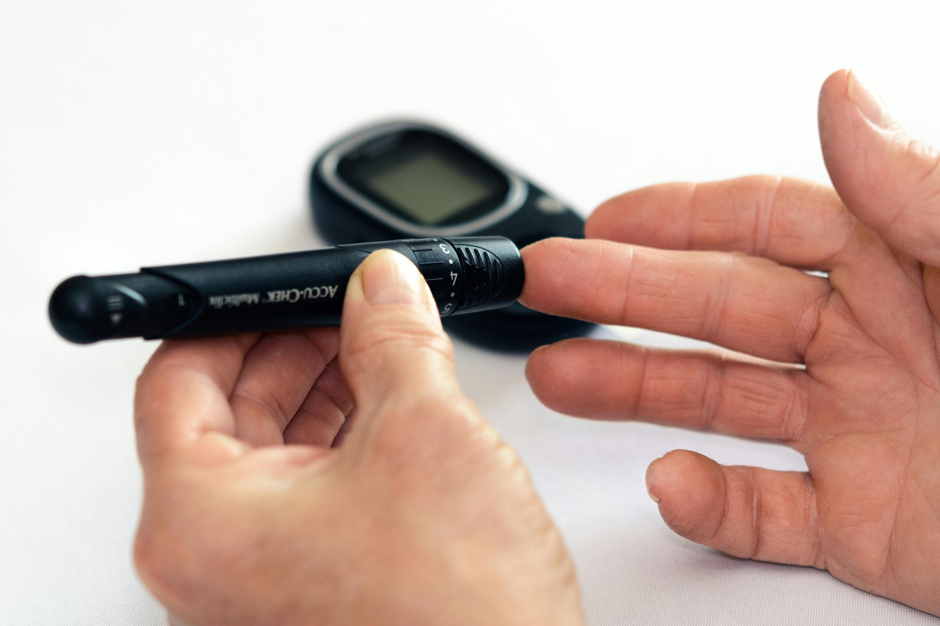 Person's hand using a lancet to prick a finger, with a blood glucose meter in the background on a white surface.