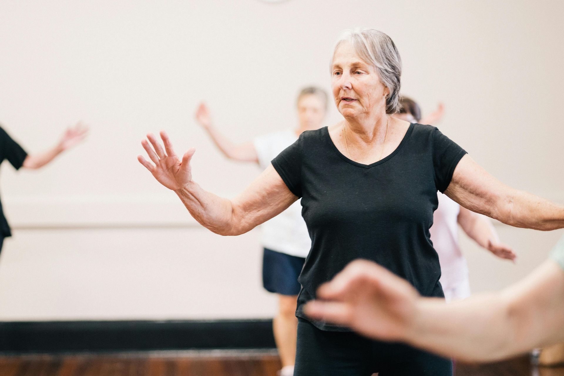 Older woman with gray hair in a black top, arms outstretched, leading a group exercise class in a studio.