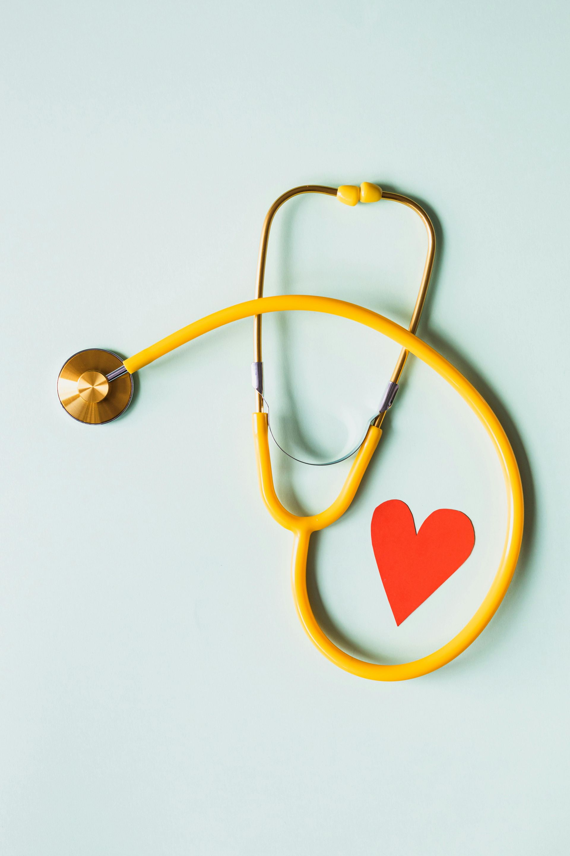 Yellow stethoscope around a red paper heart on a light blue background, representing healthcare and heart health.