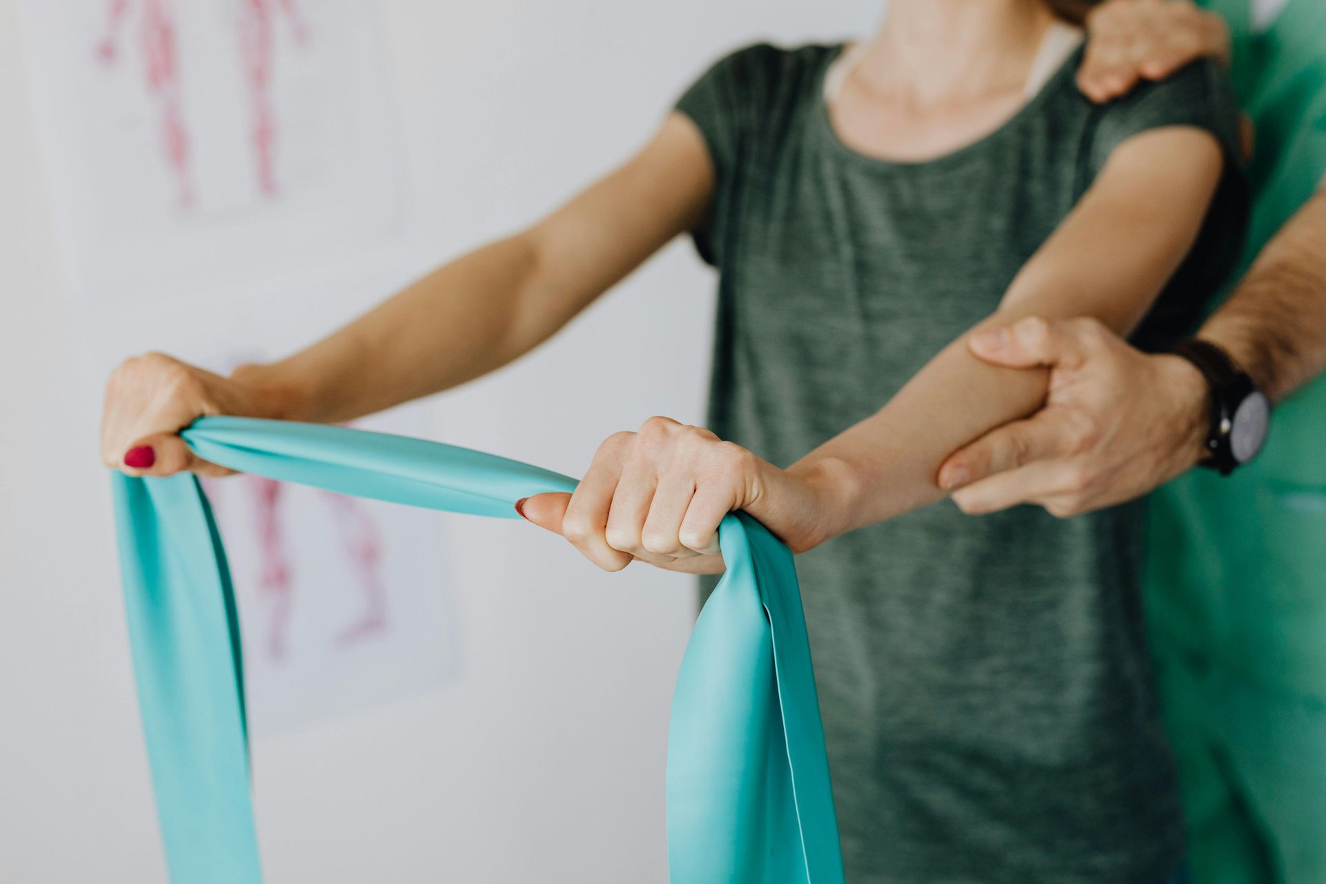 Woman in a green shirt uses a blue resistance band during physical therapy, assisted by a therapist's hands.